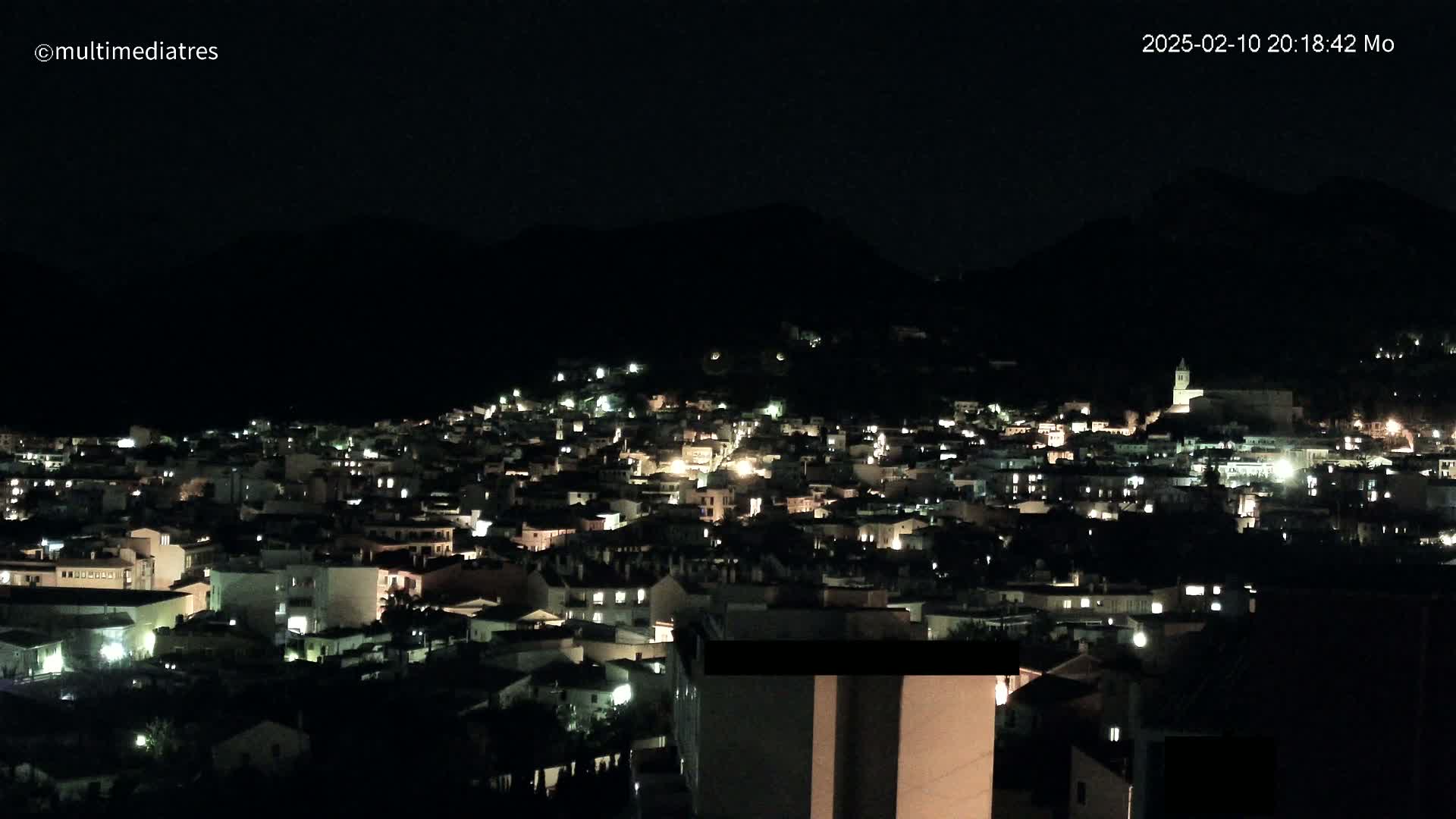 A nighttime, hilltop view of a town illuminated by scattered lights against a dark, mountainous backdrop under a clear sky.