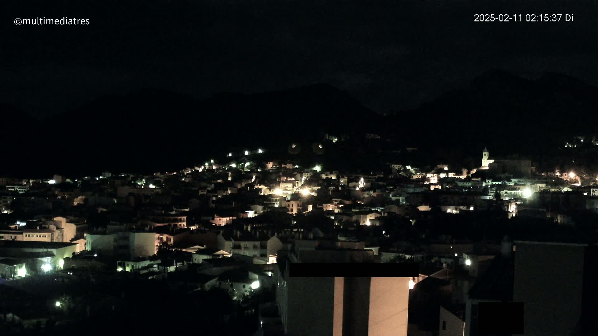 A nighttime view of a town nestled against a dark mountain, with scattered lights illuminating buildings and streets.