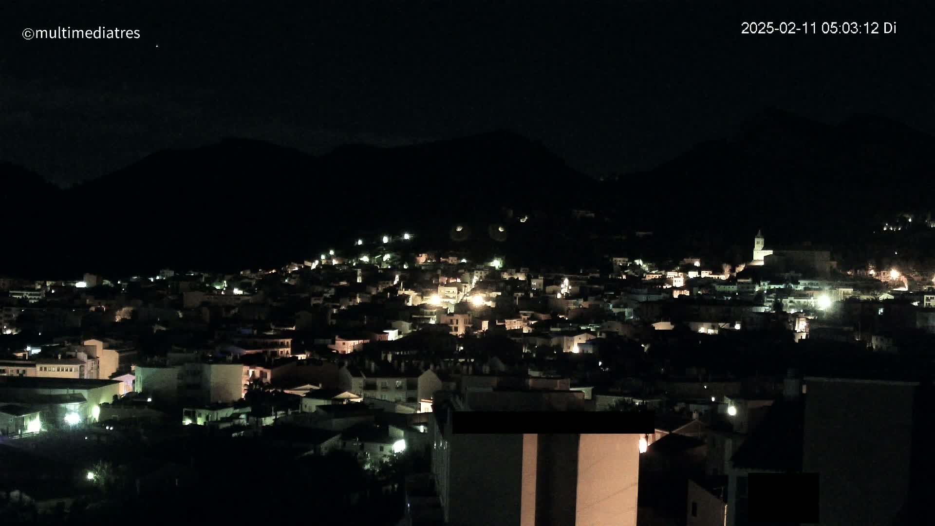 A nighttime, high-angle view of a town nestled against a dark mountain, illuminated by scattered lights.