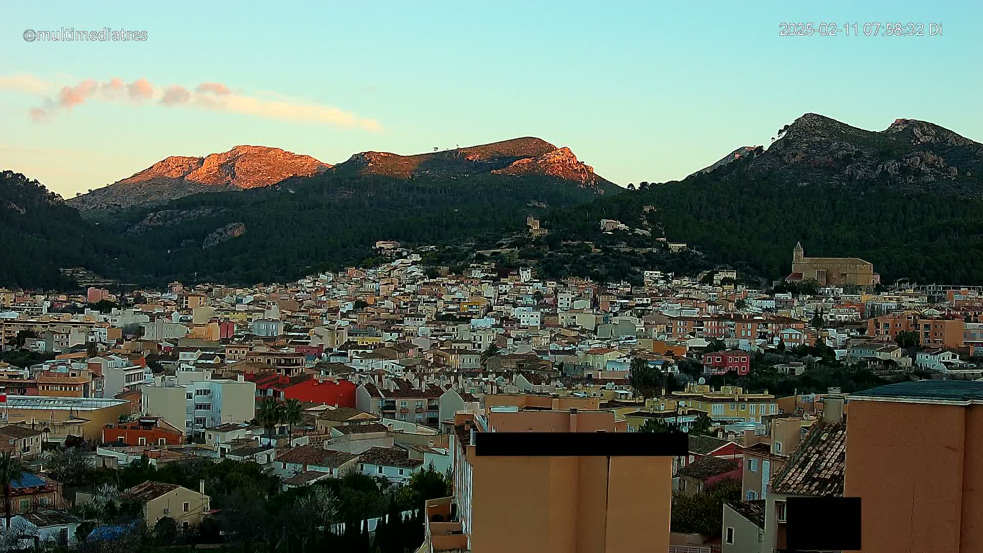 A dense collection of buildings nestled in a valley, with sunlit mountains in the background under a clear, twilight sky.