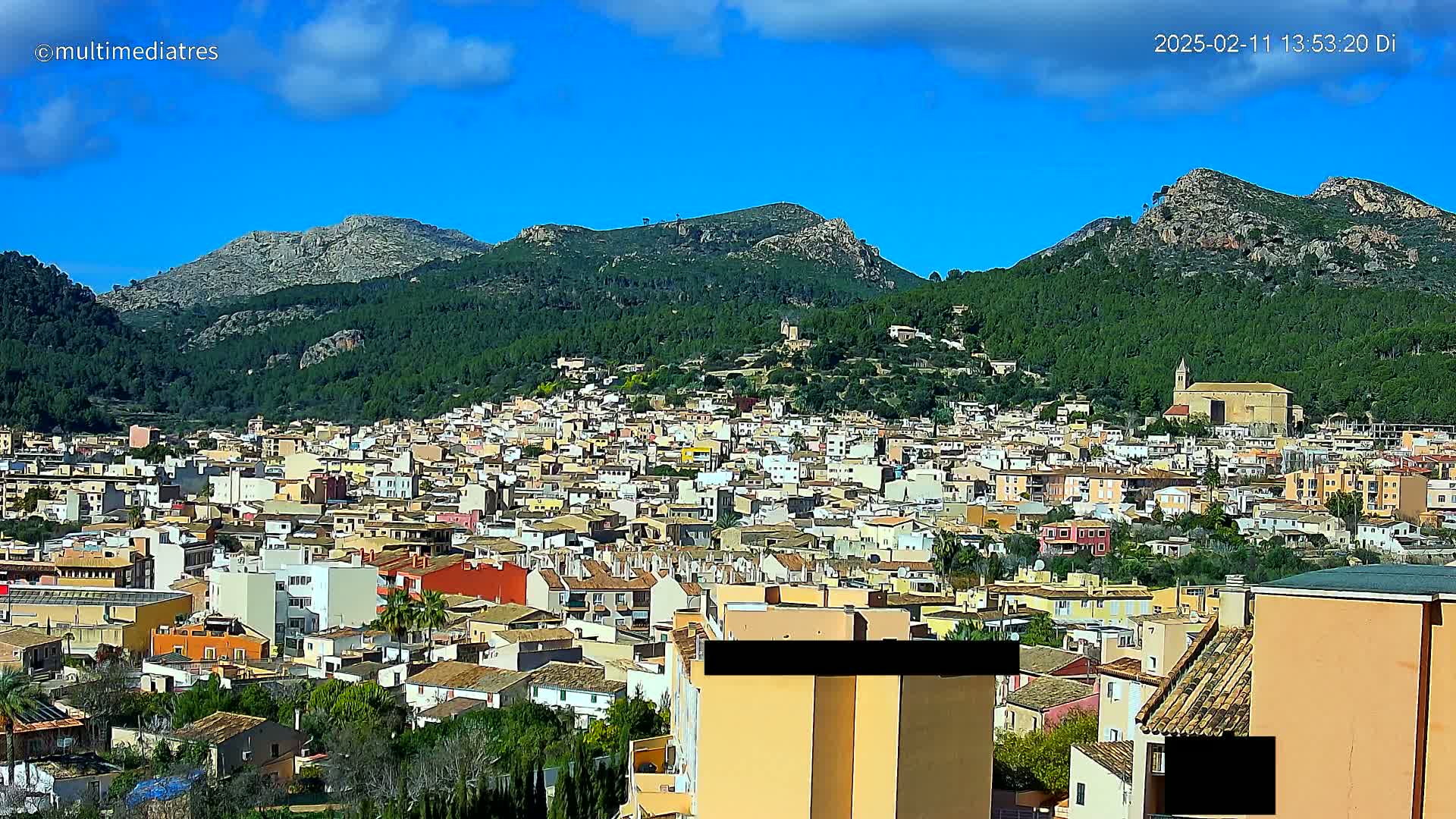 A sprawling town of predominantly low-rise buildings nestles at the foot of verdant, rocky mountains under a mostly sunny sky with a few scattered clouds.