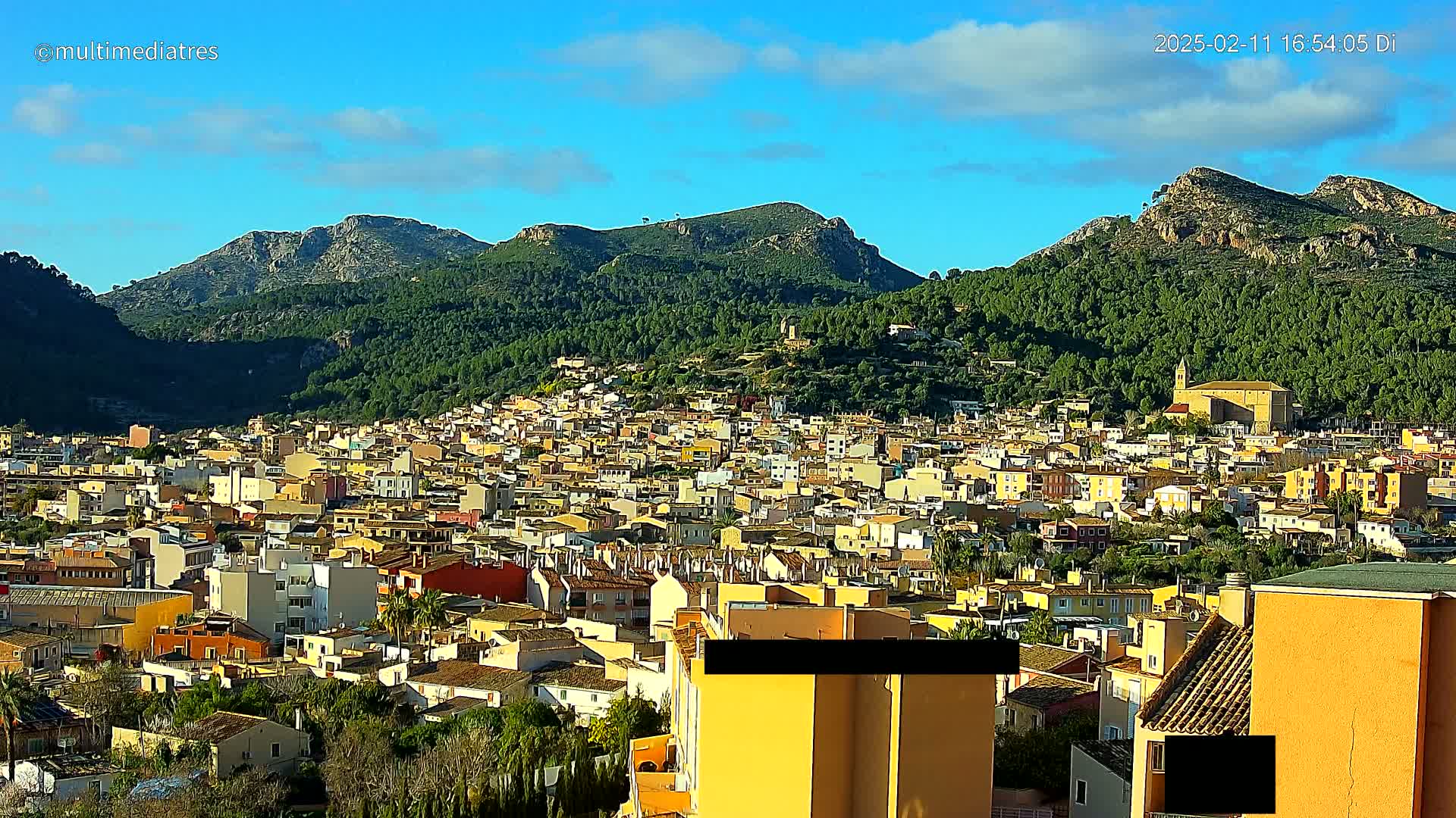 A sprawling town of light-colored buildings nestles at the foot of lush green mountains under a mostly clear, bright blue sky.