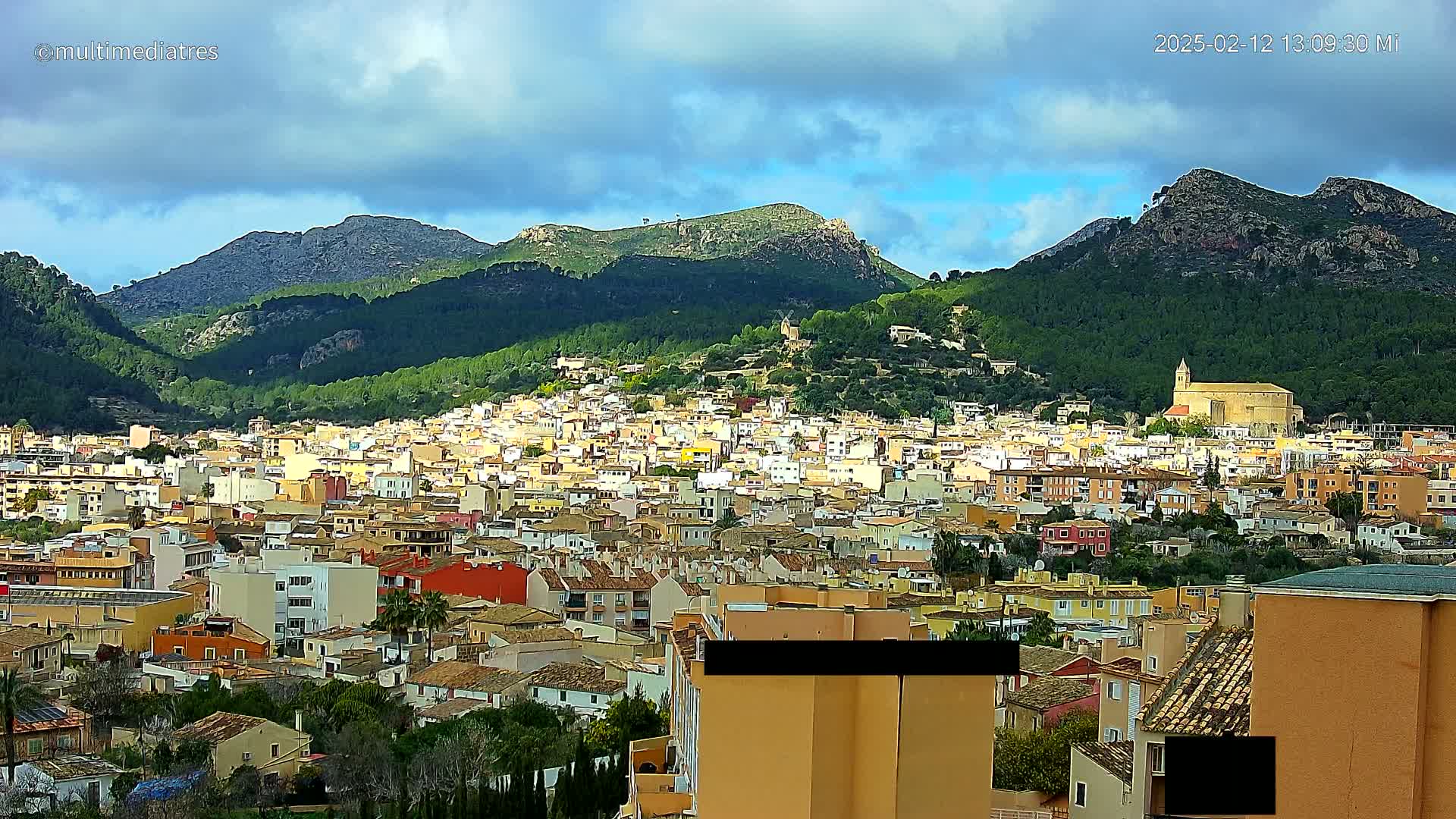 A sprawling town of low-rise buildings nestles at the base of verdant, mountainous terrain under a partly cloudy sky.