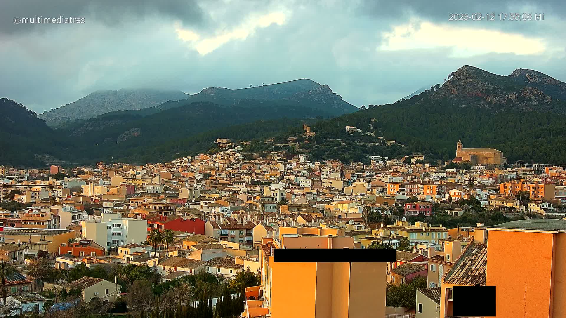 A sprawling town of tan and orange buildings nestled in a valley between verdant mountains under a cloudy sky.