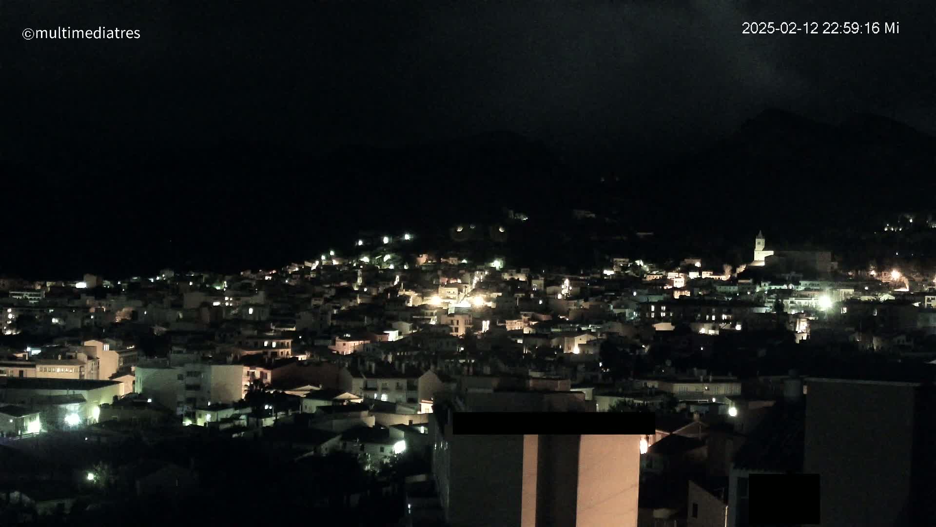 A nighttime view of a town nestled in a dark hillside, illuminated by scattered lights.