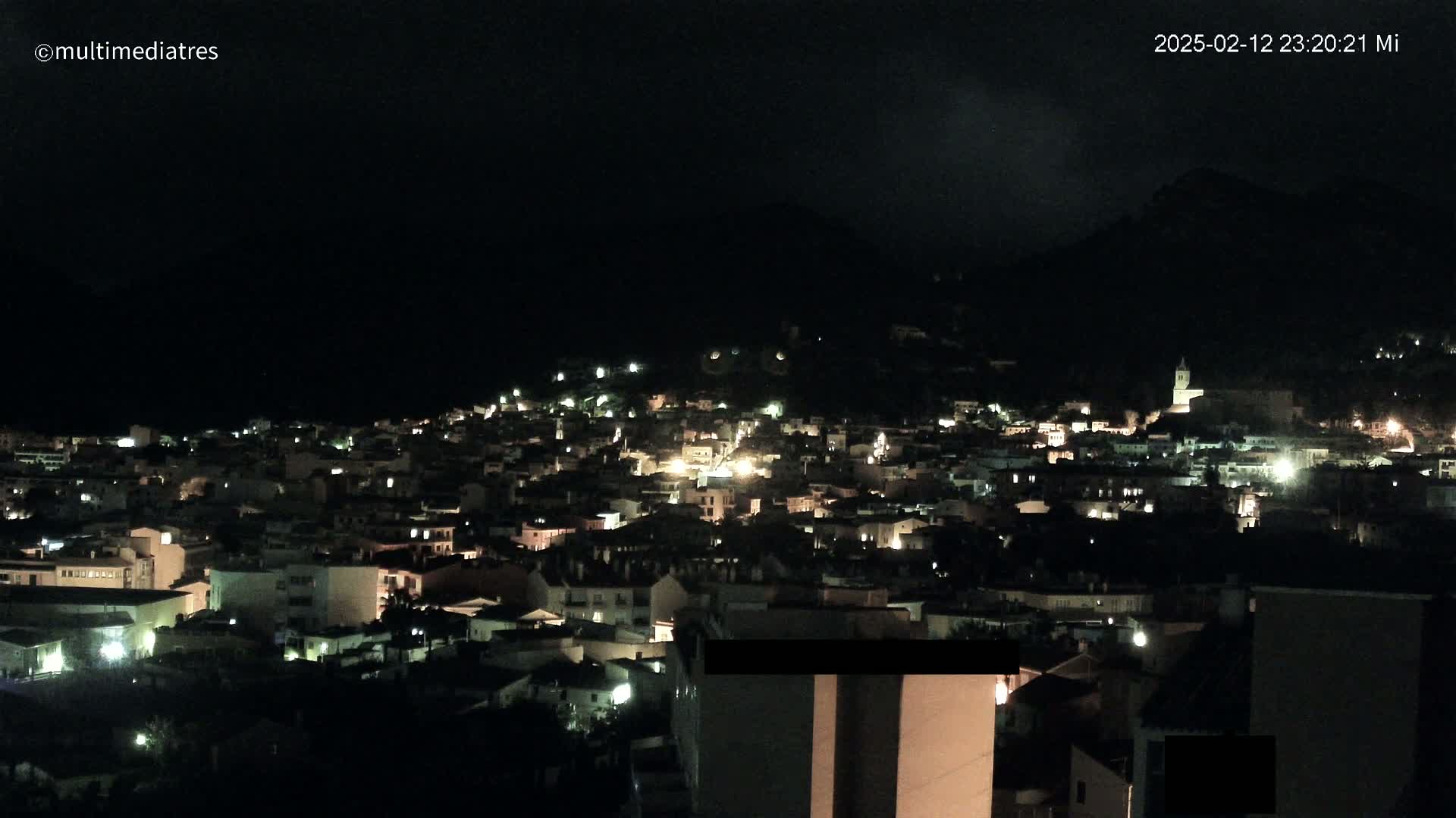 A nighttime, aerial view shows a town nestled against a dark mountain, with scattered lights illuminating the buildings below.