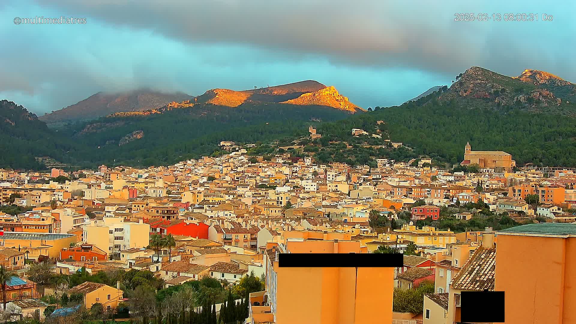 A sprawling town of predominantly orange and yellow buildings nestled in a valley between mountains, under a partly cloudy sky with sunlight illuminating the highest peaks.