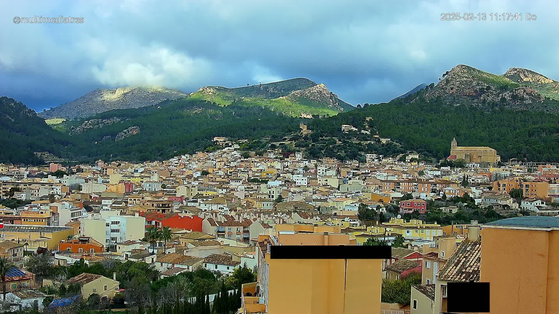 A sprawling town of colorful buildings nestles at the base of verdant, rocky mountains under a partly cloudy sky.