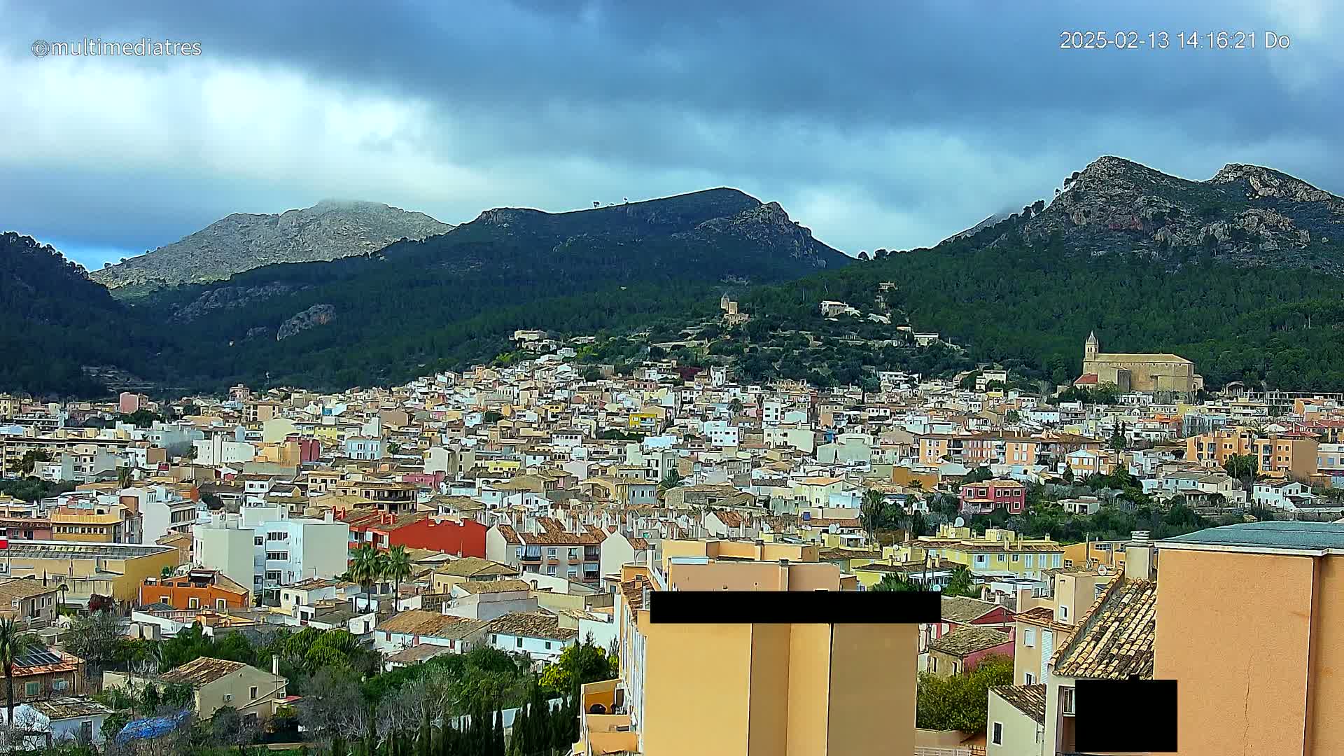 A sprawling town of colorful buildings nestles in a valley surrounded by verdant, mountainous terrain under a mostly cloudy sky.
