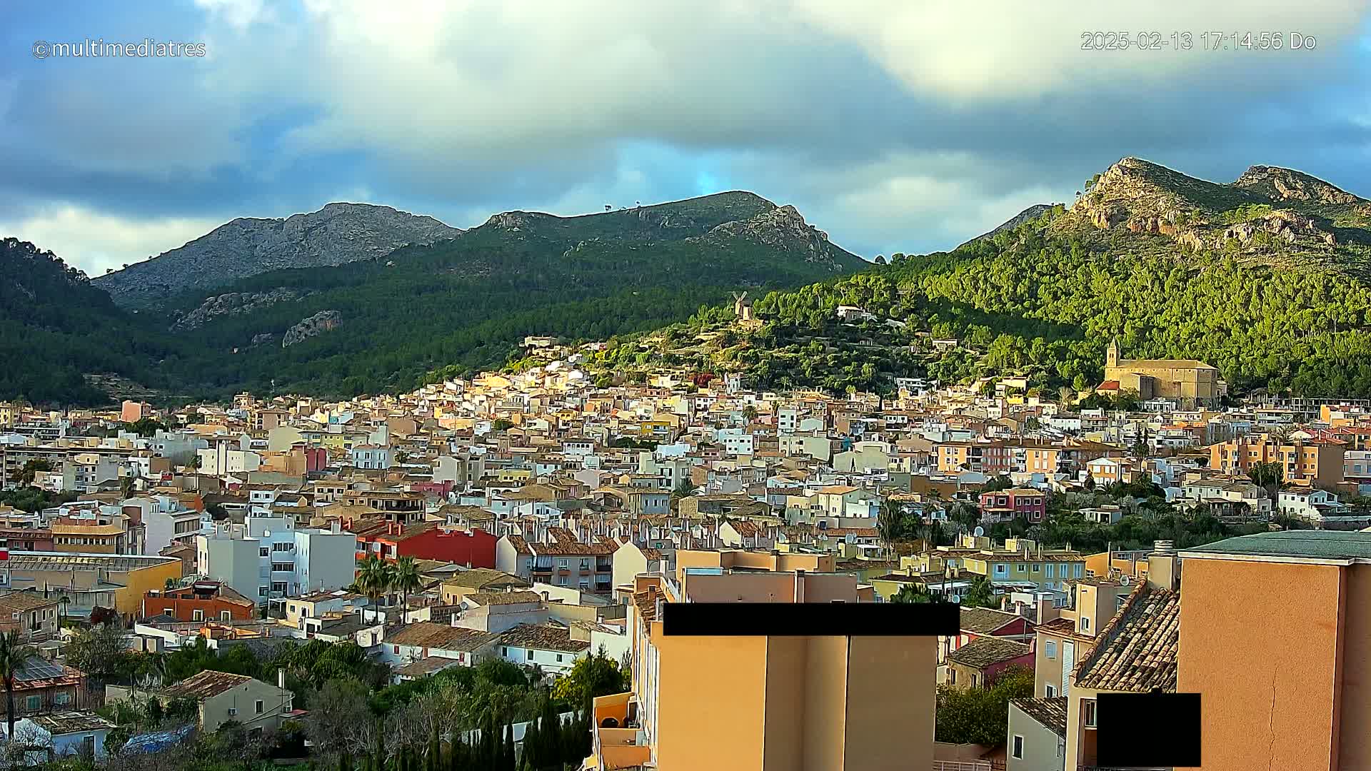 A sprawling town of closely packed buildings nestles in a valley between verdant, rocky hills under a partly cloudy sky.