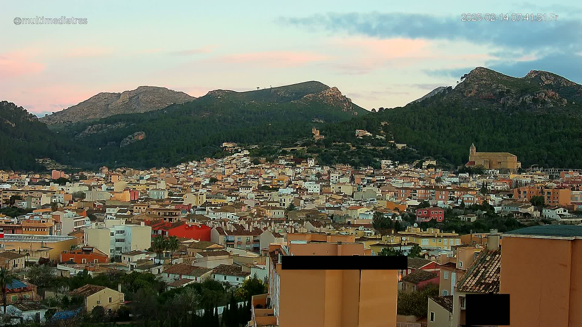 A sprawling town of pastel-colored buildings nestles at the foot of verdant, mountainous terrain under a soft, pinkish sunset sky.