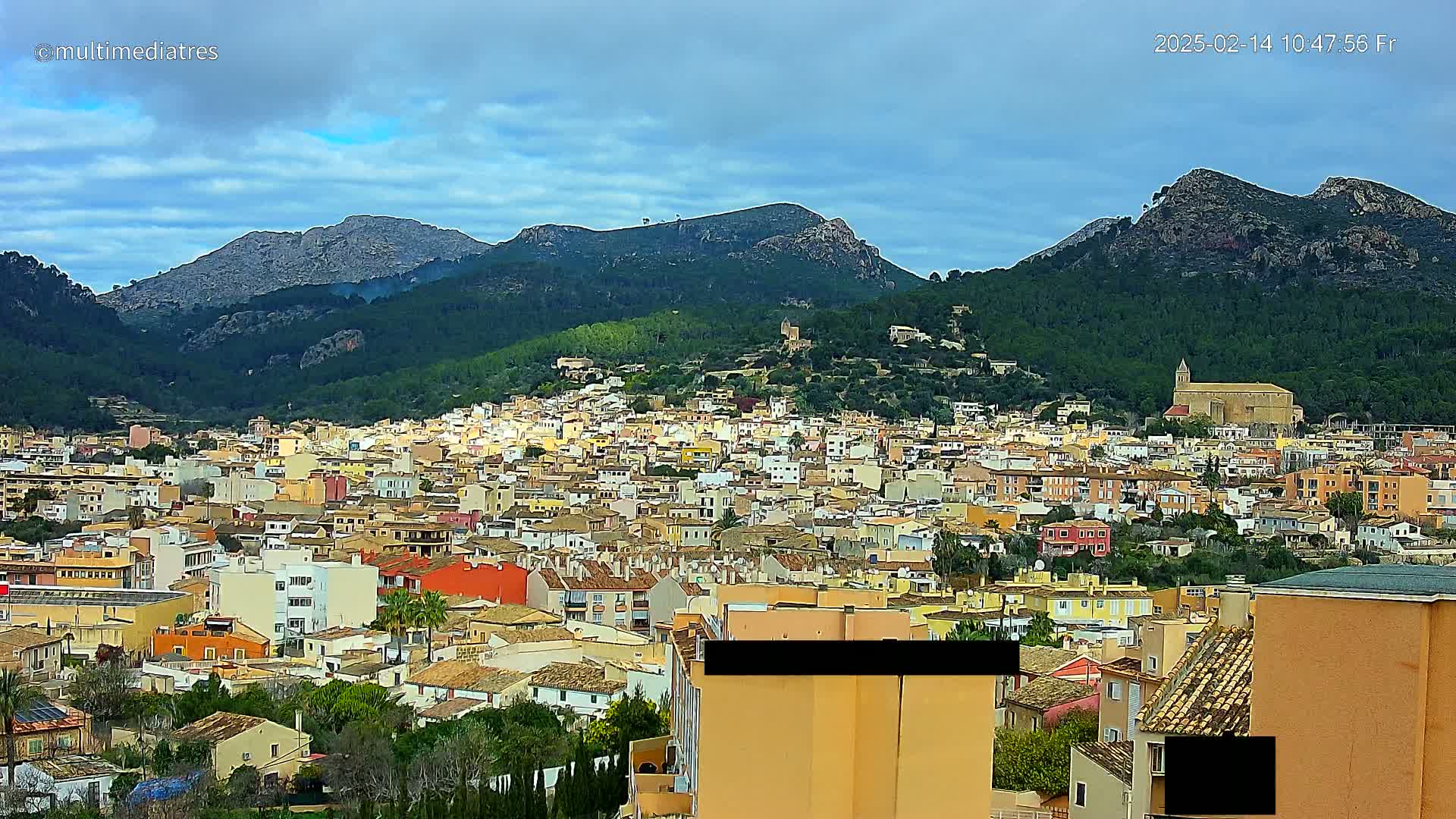 A sprawling town of tan and orange buildings nestles at the base of a mountain range under a partly cloudy sky.