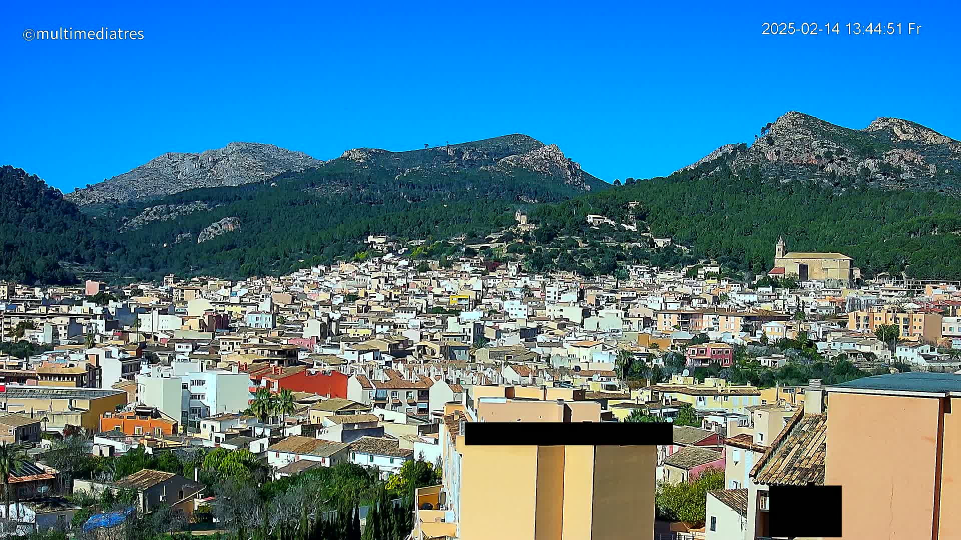 A sprawling town of tan and orange buildings nestled at the base of verdant, rocky mountains under a clear blue sky.