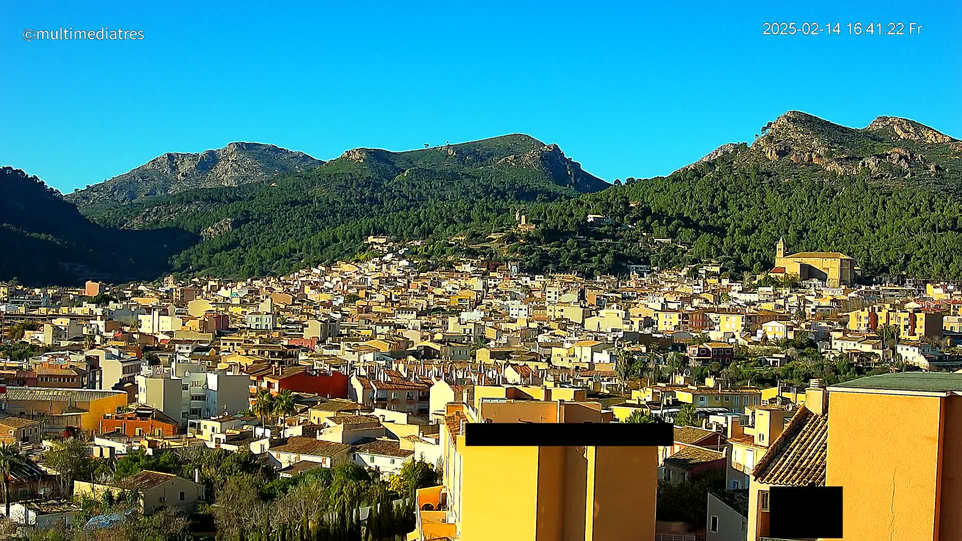 A sprawling town of predominantly low-rise, light-colored buildings nestles at the base of sunlit, verdant mountains under a clear blue sky.