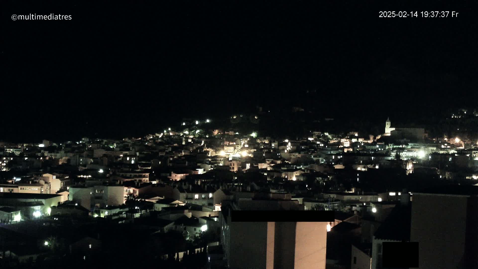 A nighttime, aerial view of a town nestled on a hillside, illuminated by scattered lights.