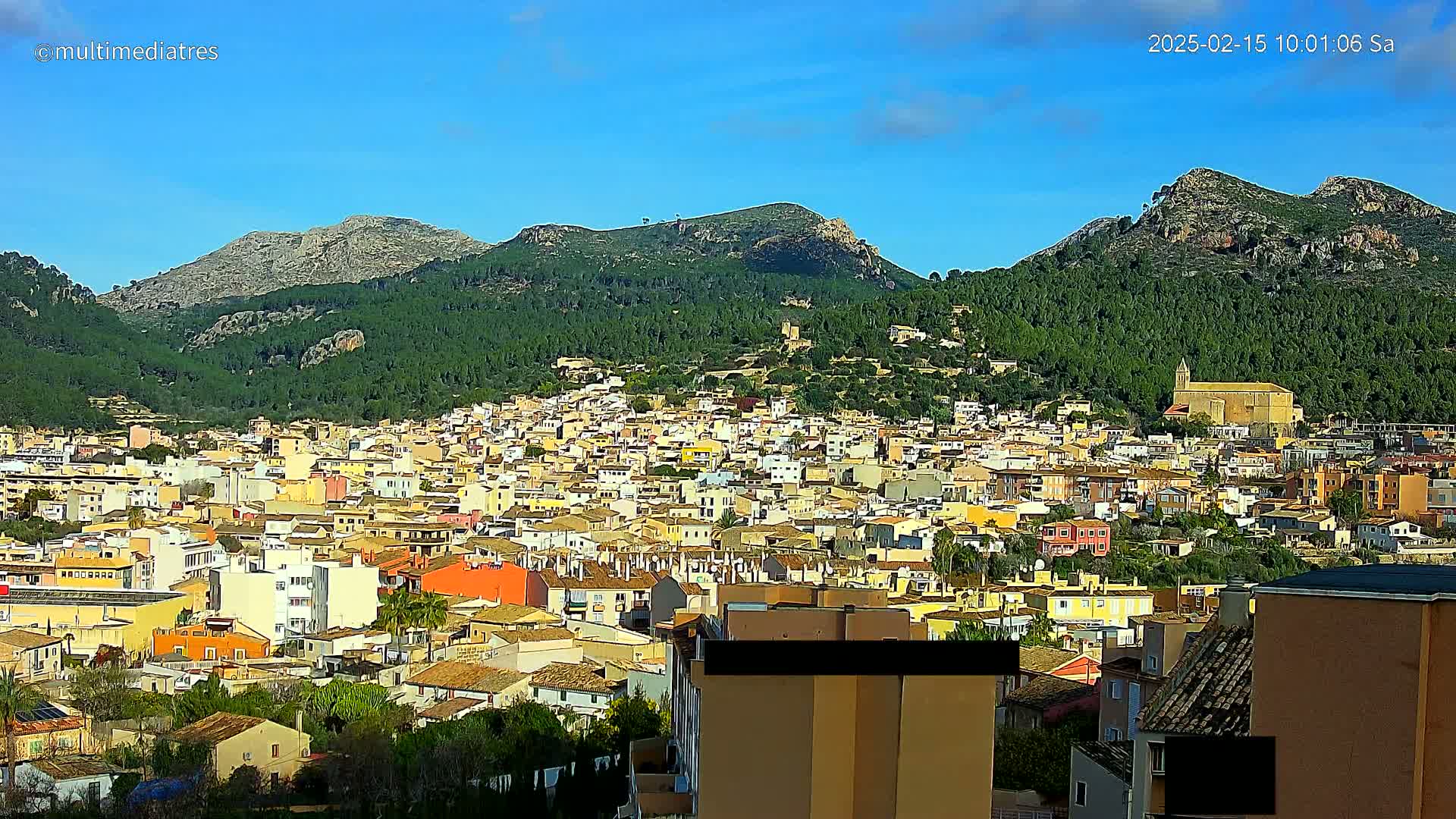 A sprawling town of tan and yellow buildings nestled in a verdant valley, backed by sunlit, rocky mountains under a clear blue sky.