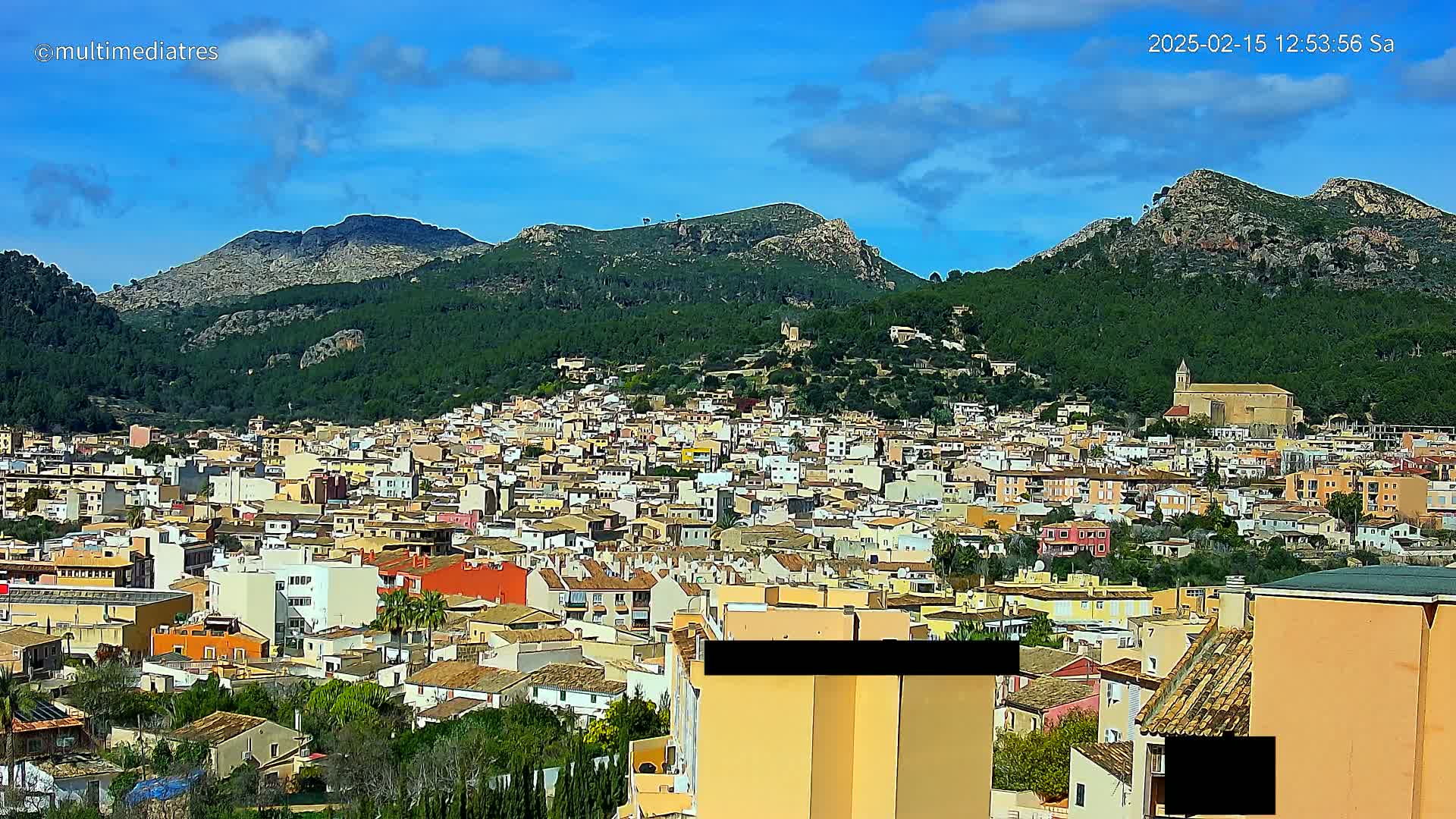 A sprawling town of light-colored buildings nestled at the base of verdant, rocky mountains under a mostly clear blue sky.