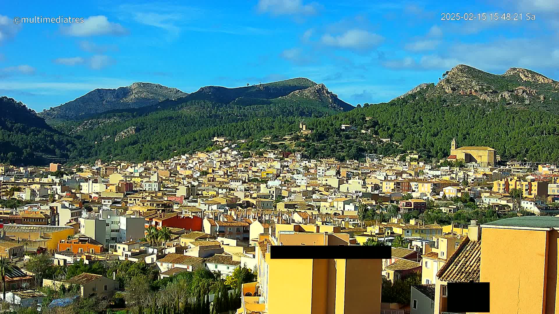 A dense collection of buildings nestled at the foot of a lush, mountainous landscape under a bright, sunny sky.