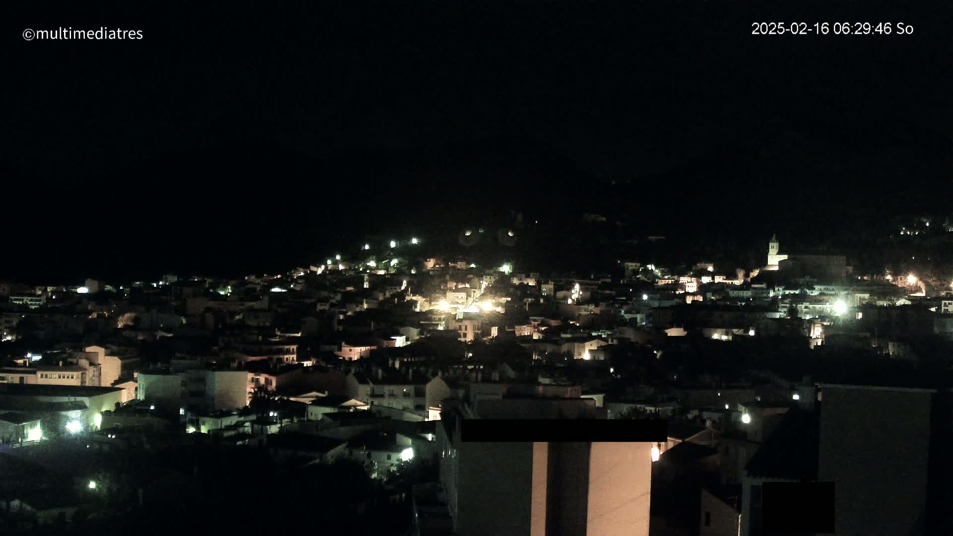 A nighttime, aerial view of a town nestled on a hill, illuminated by scattered lights.