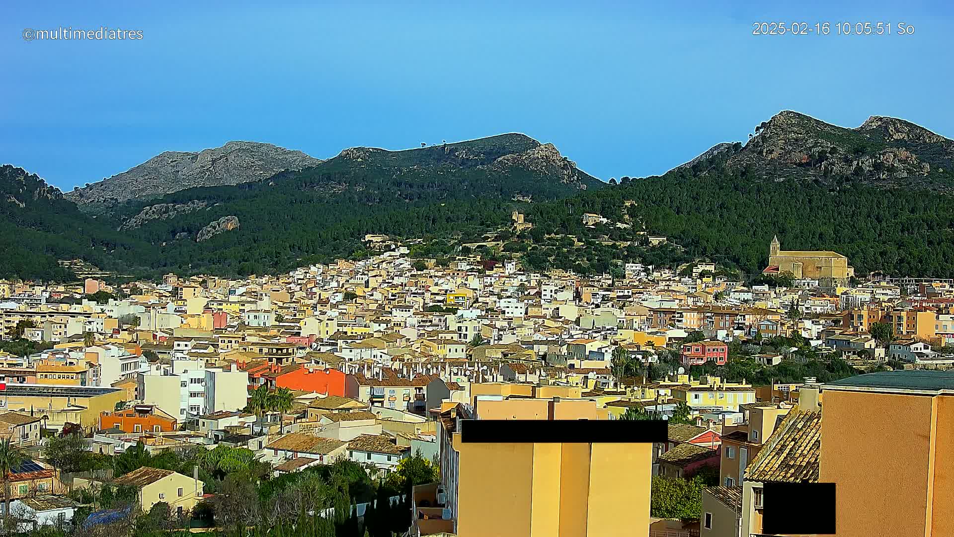 A sprawling town of low-rise buildings, nestled in a valley surrounded by green, mountainous terrain under a clear blue sky.