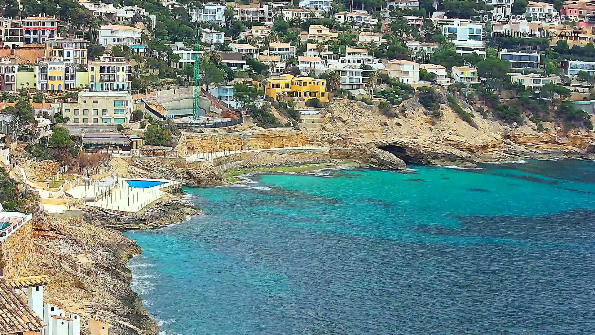 A coastal area with a cluster of buildings on a hillside overlooking a turquoise bay with a small swimming pool and walkway next to the water.