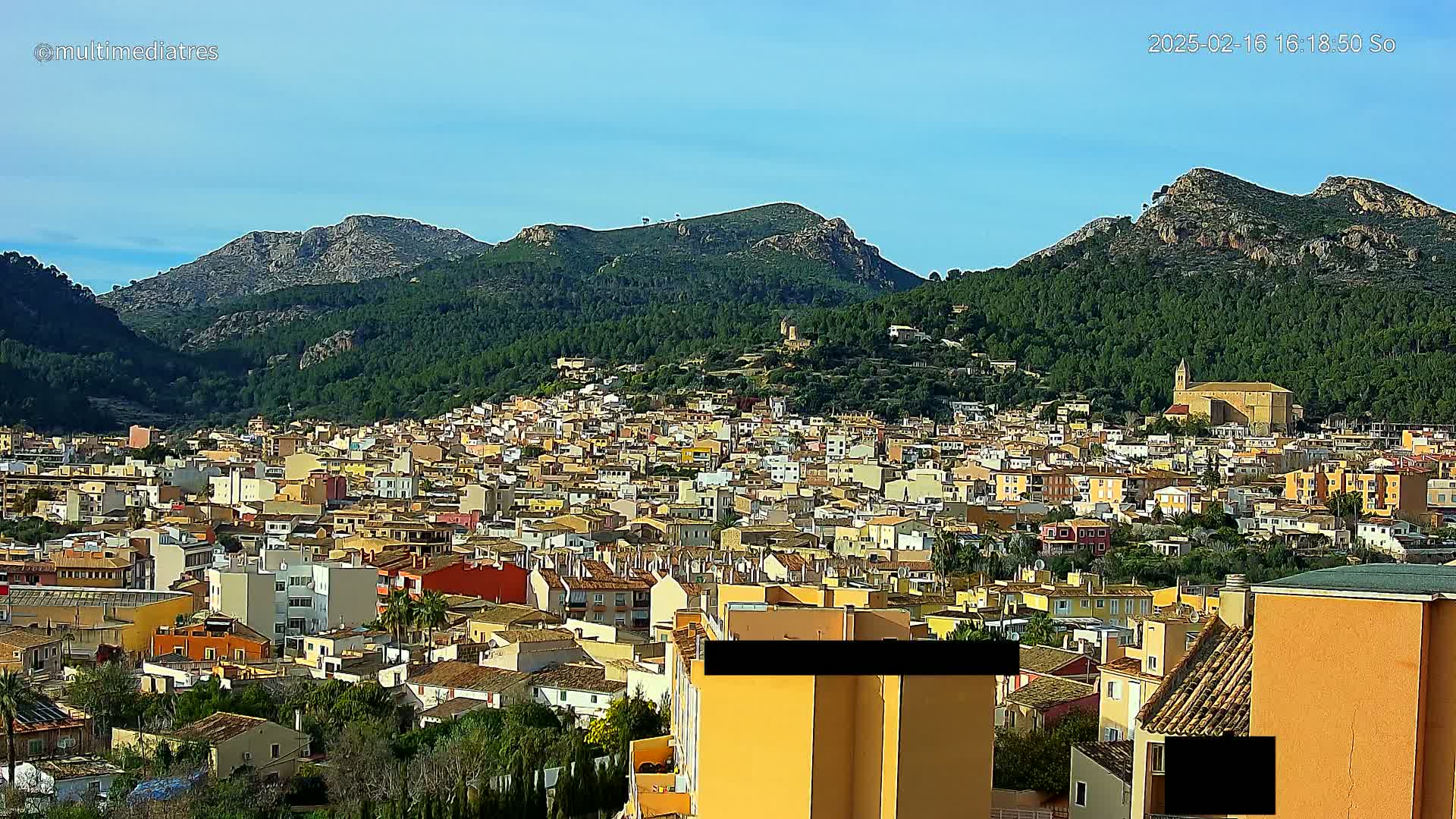 A dense cluster of low-rise buildings in a town nestled in a valley surrounded by green, mountainous terrain under a clear, bright sky.