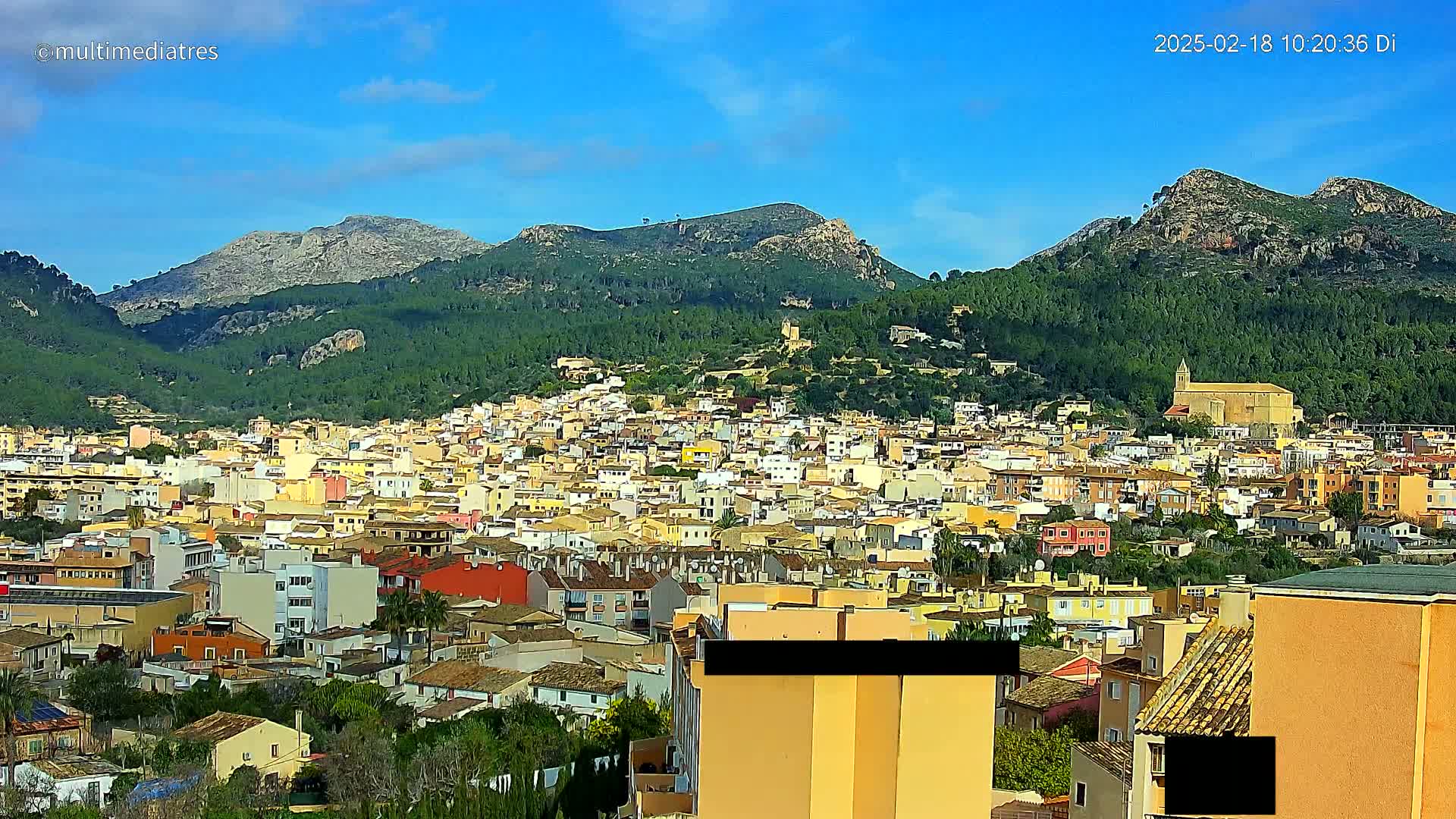 A sprawling town of tan and yellow buildings nestles in a valley between green, mountainous hills under a clear blue sky.