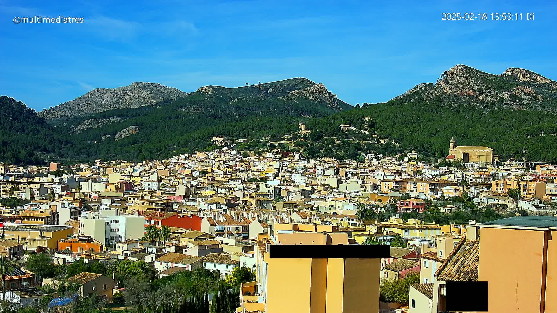 A sprawling town of low-rise buildings nestles in a valley between verdant, rocky mountains under a clear blue sky.