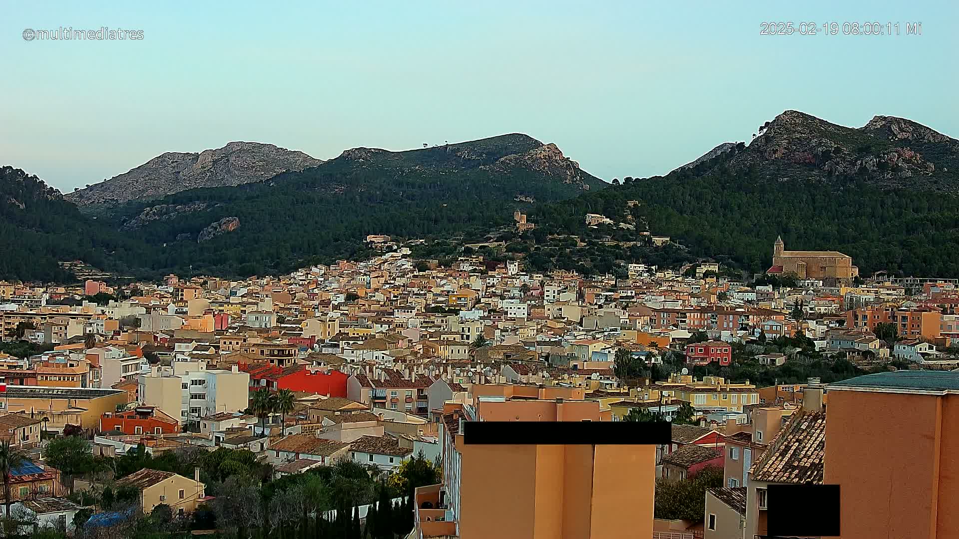A sprawling town of low-rise buildings nestles at the base of a verdant, mountainous region under a clear, light-blue sky.