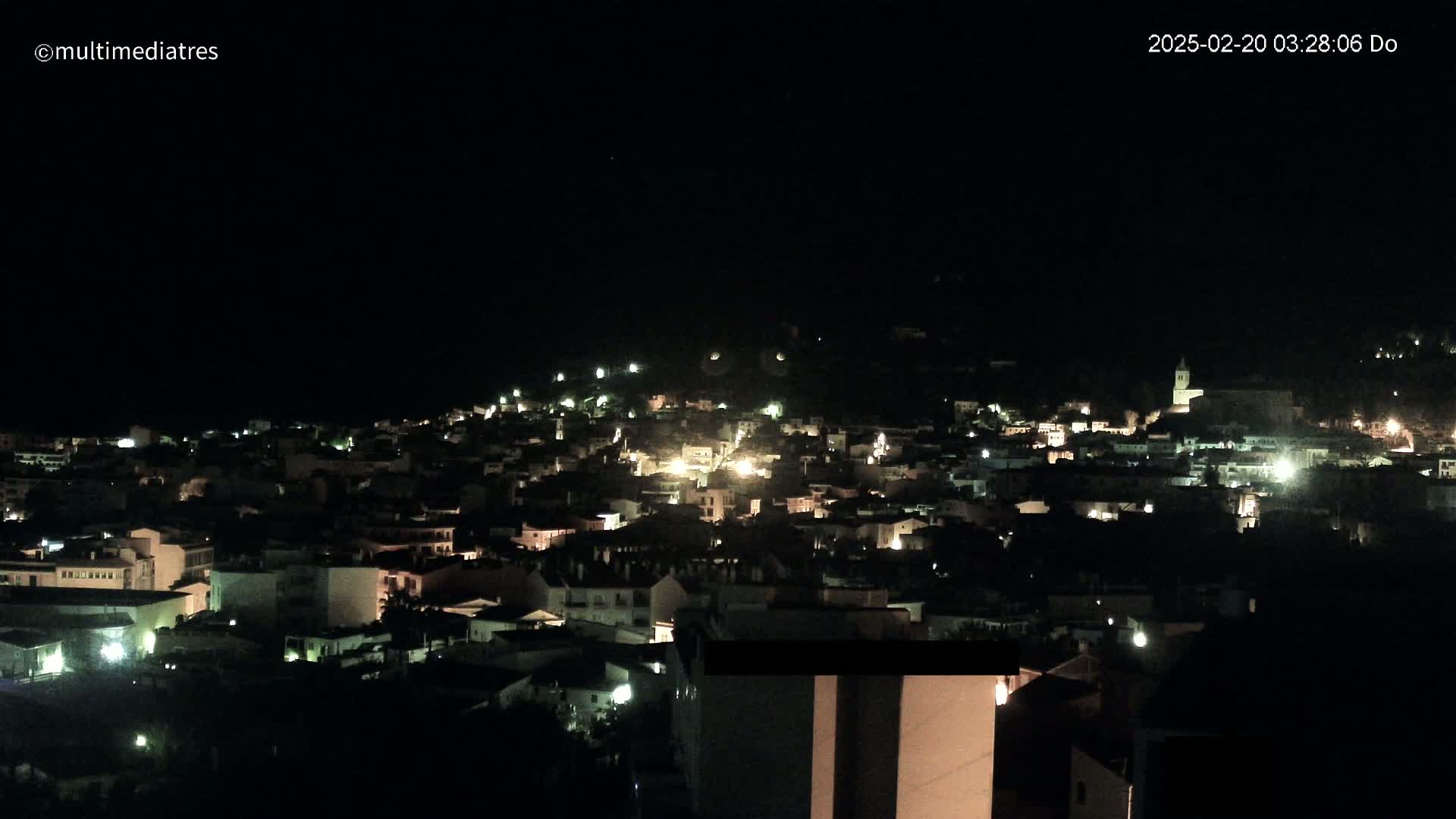 A nighttime view of a town nestled on a hillside, illuminated by scattered lights under a clear, dark sky.