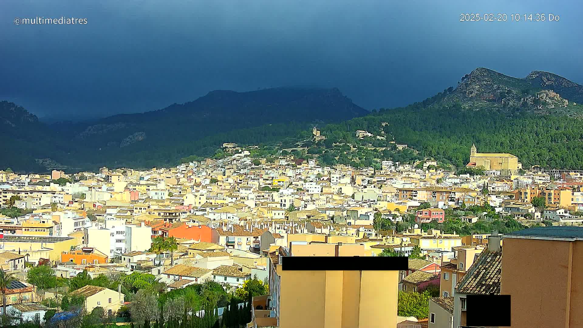 A sprawling town of low-rise buildings nestles in a valley, overlooked by dark, storm-laden skies and verdant hills.