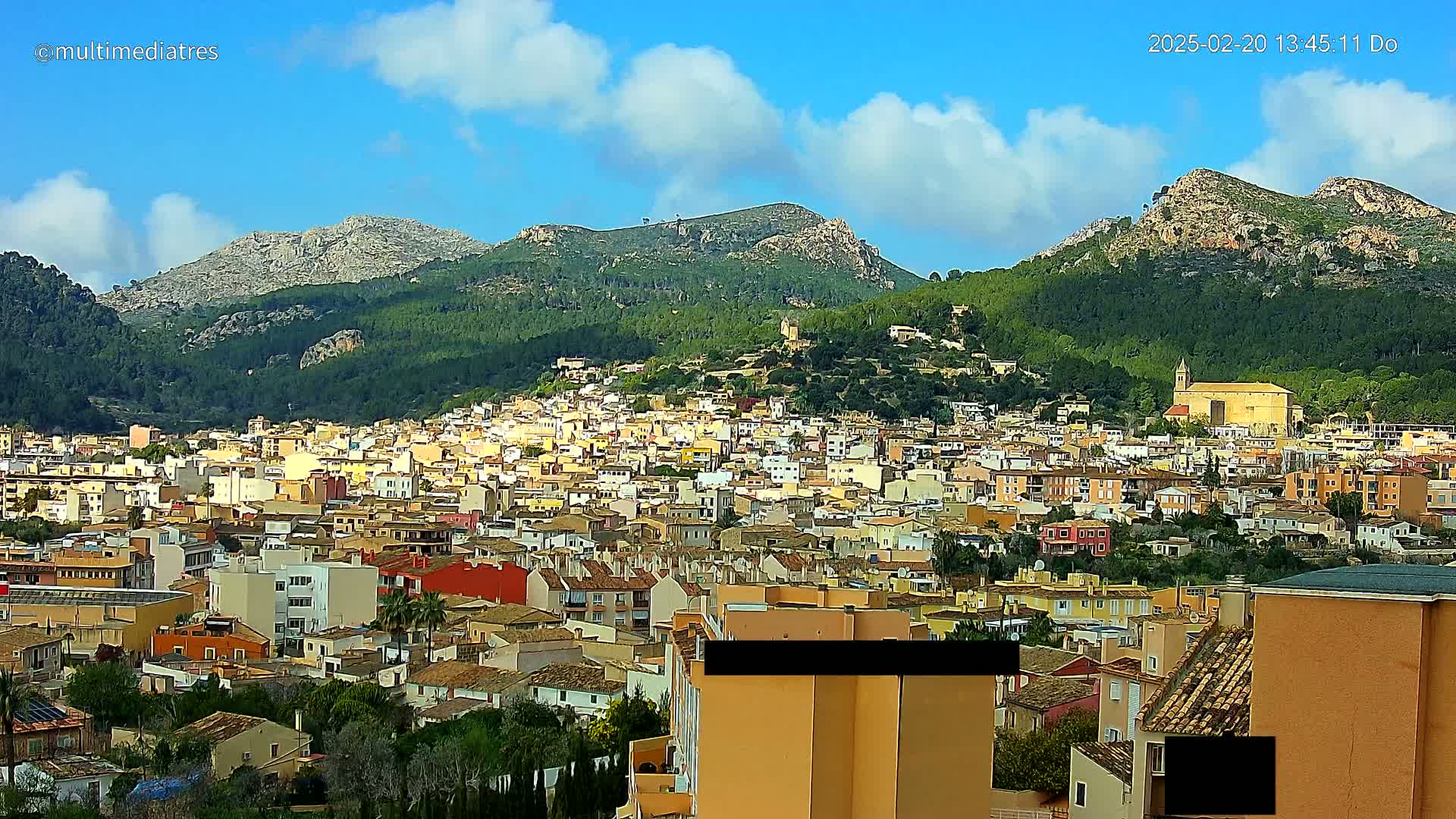 A sprawling town of low-rise buildings nestles in a valley between green, tree-covered mountains under a partly cloudy blue sky.