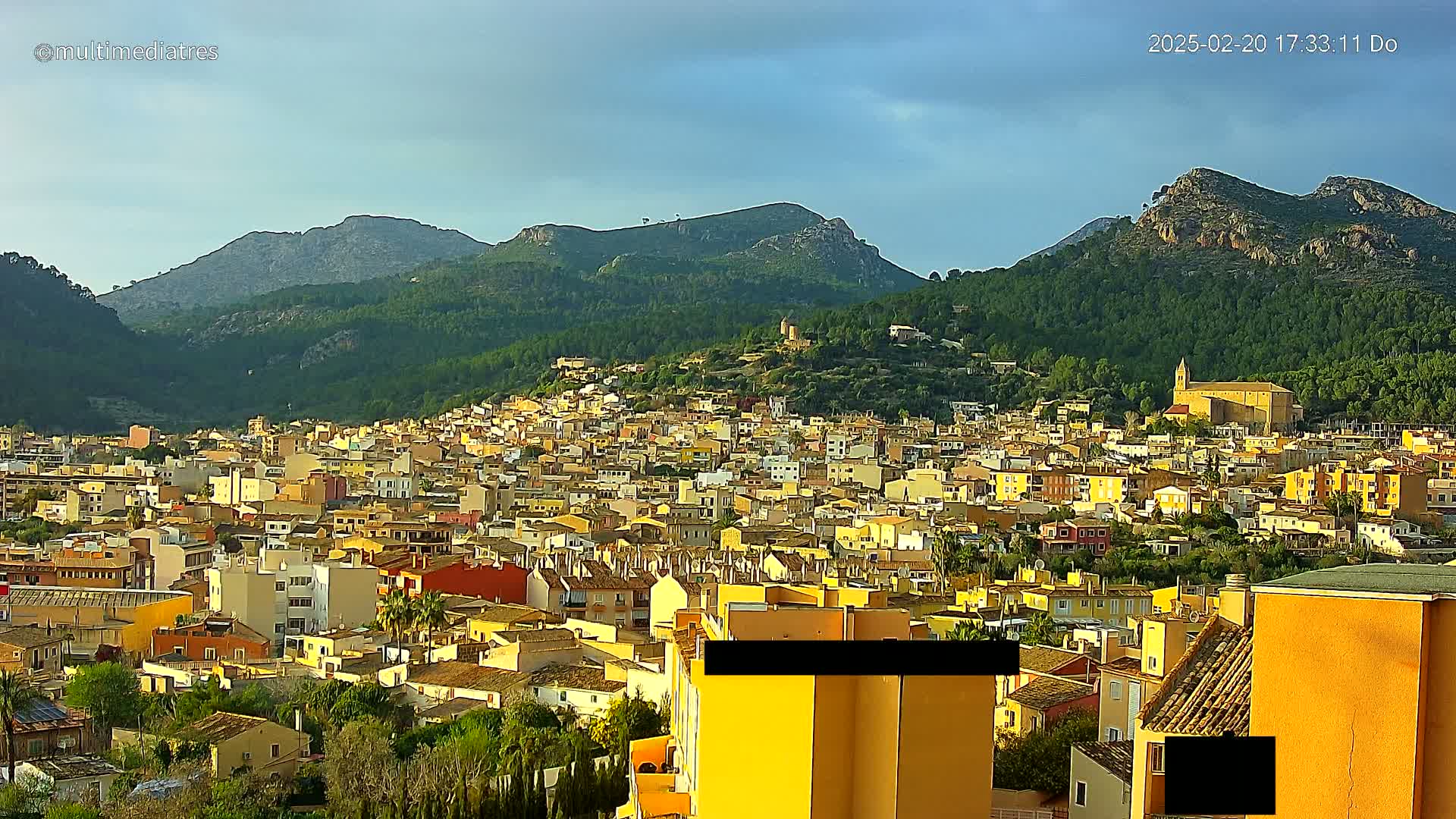 A sprawling town of yellow and orange buildings nestles in a valley surrounded by green, mountainous terrain under a partly cloudy sky.