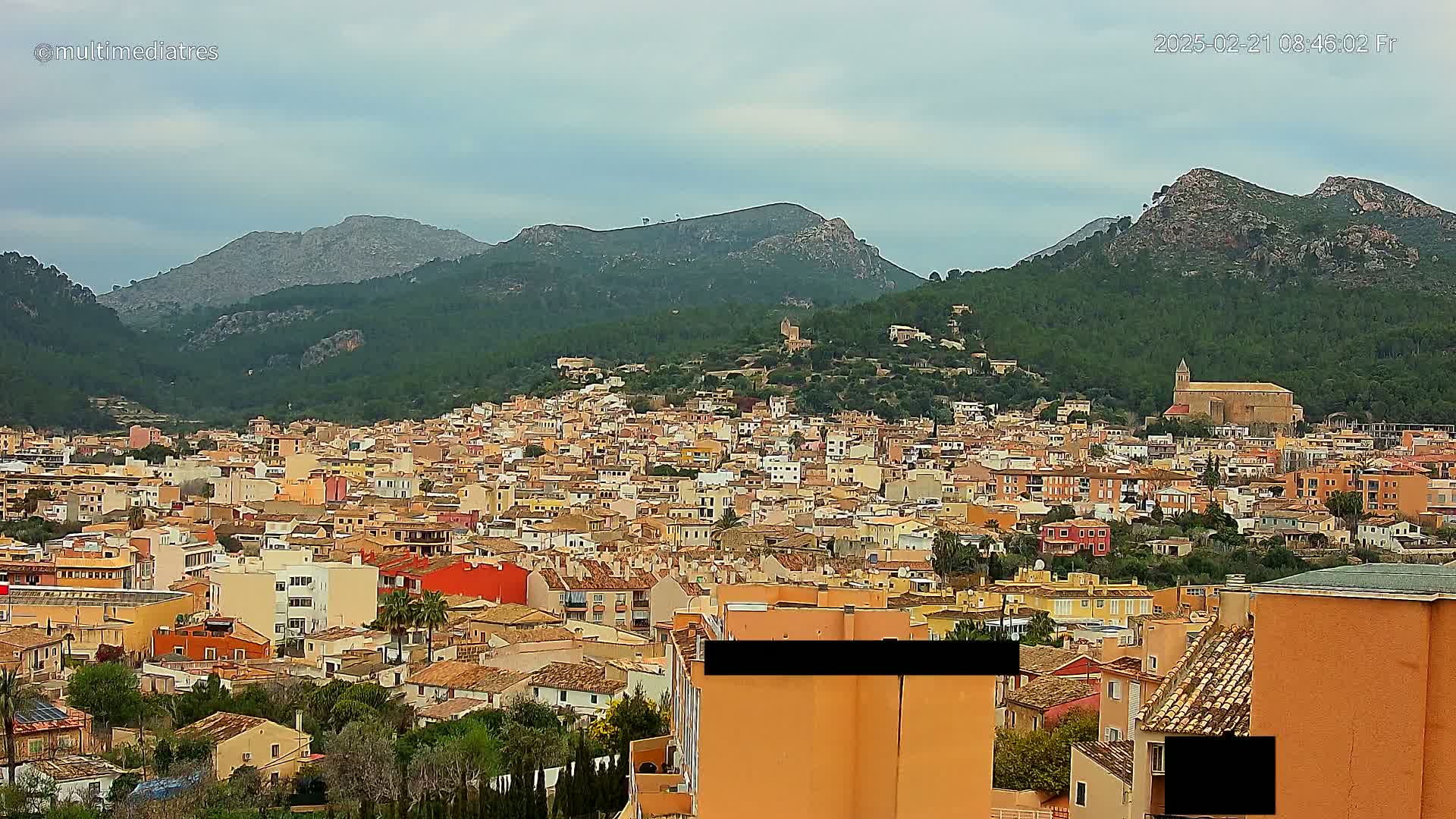A sprawling town of low-rise buildings with orange roofs nestles in a green valley surrounded by mountains under a cloudy sky.