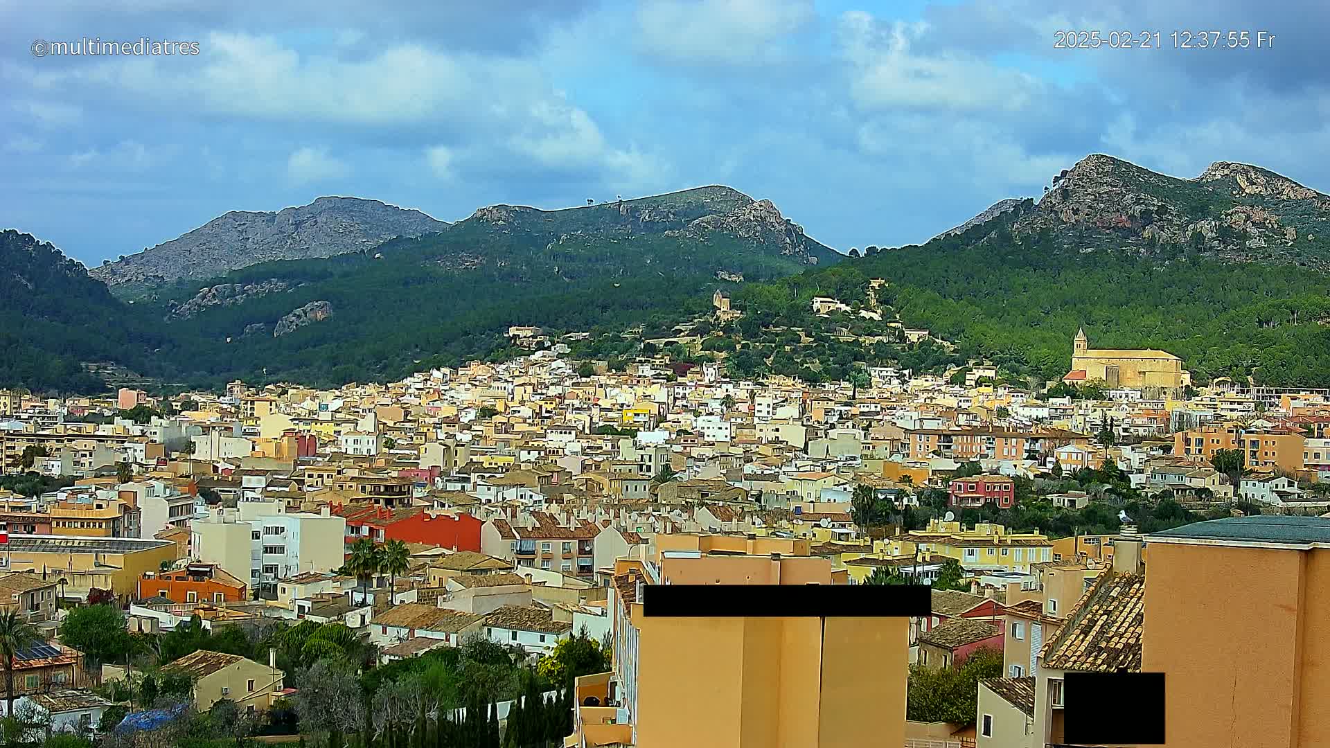 A sprawling town of low-rise buildings nestles at the foot of verdant, rocky mountains under a partly cloudy sky.