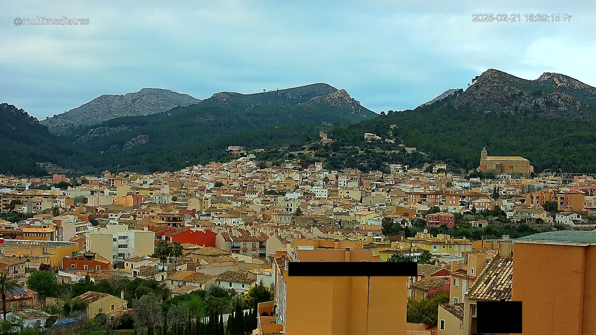 A dense cluster of buildings, mostly low-rise, stretches across a hillside, nestled beneath mountains under a cloudy sky.