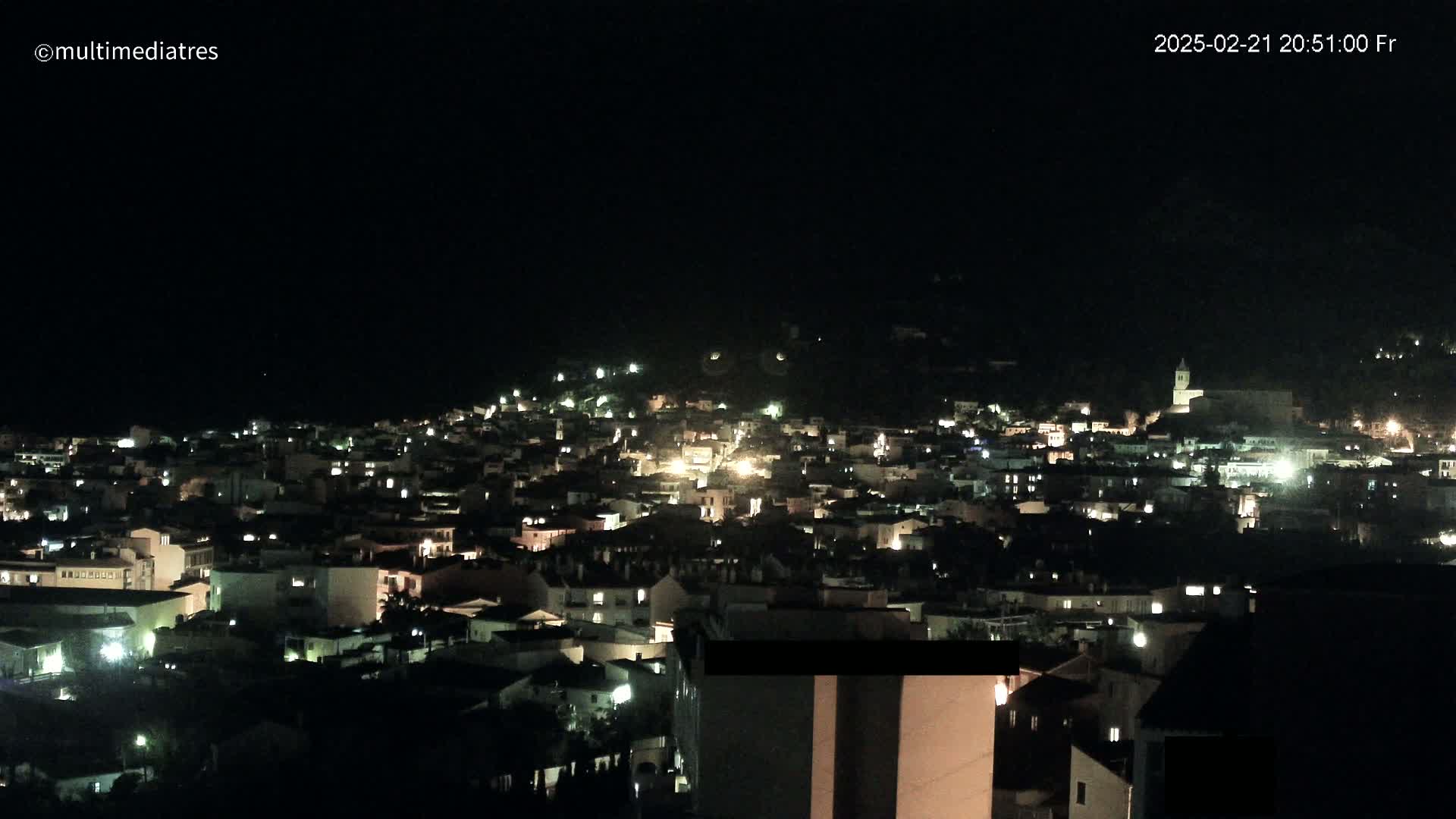 A nighttime, aerial view shows a town illuminated by scattered house lights, nestled on a dark hillside under a clear, black sky.