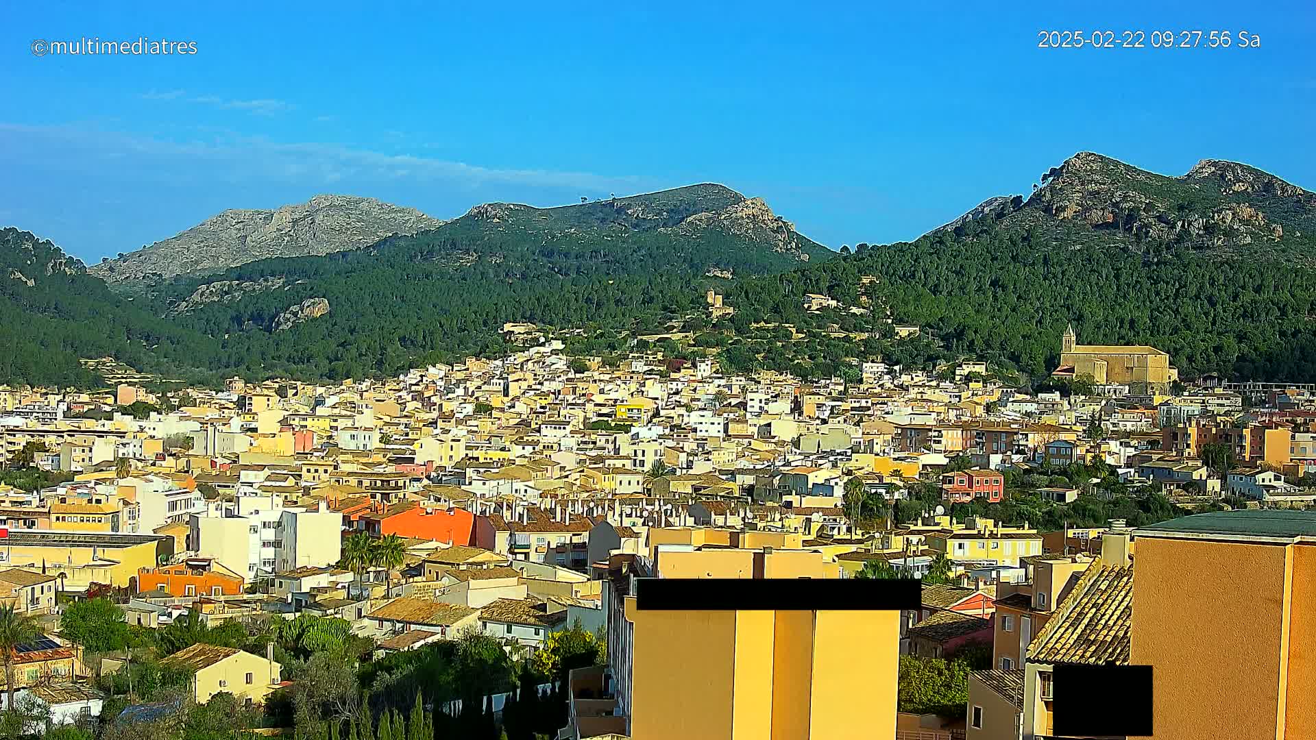 A sprawling town of low-rise buildings climbs a hillside, nestled beneath verdant mountains under a clear blue sky.