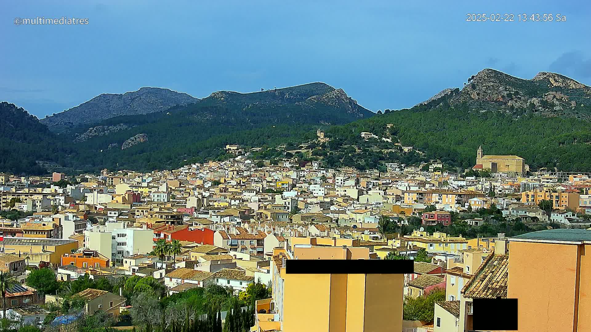 A sprawling town of low-rise buildings nestled in a valley between verdant, mountainous hills under a mostly clear sky.