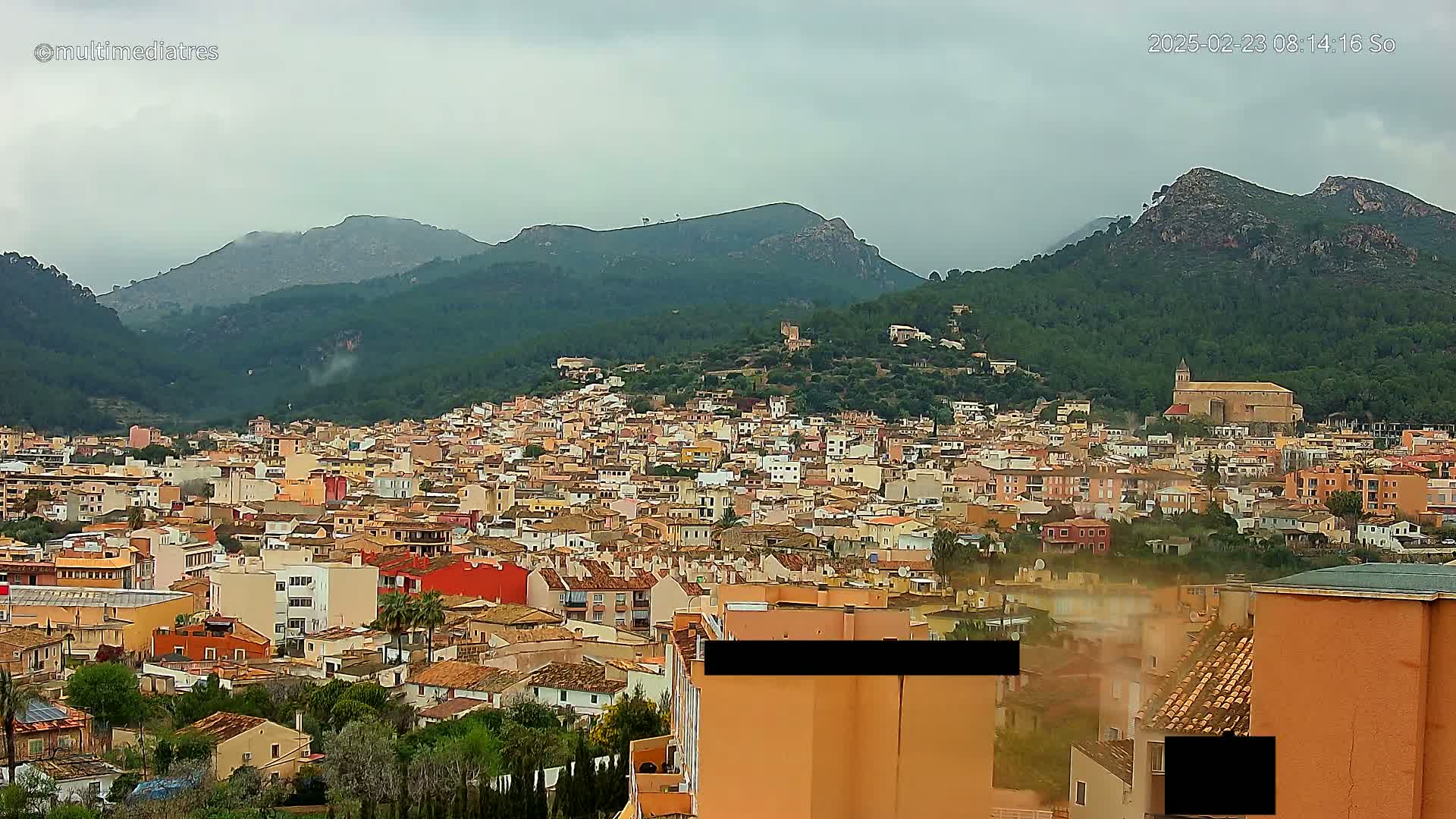 A sprawling town of tan and orange buildings nestled in a valley between verdant, mountainous terrain under an overcast sky.