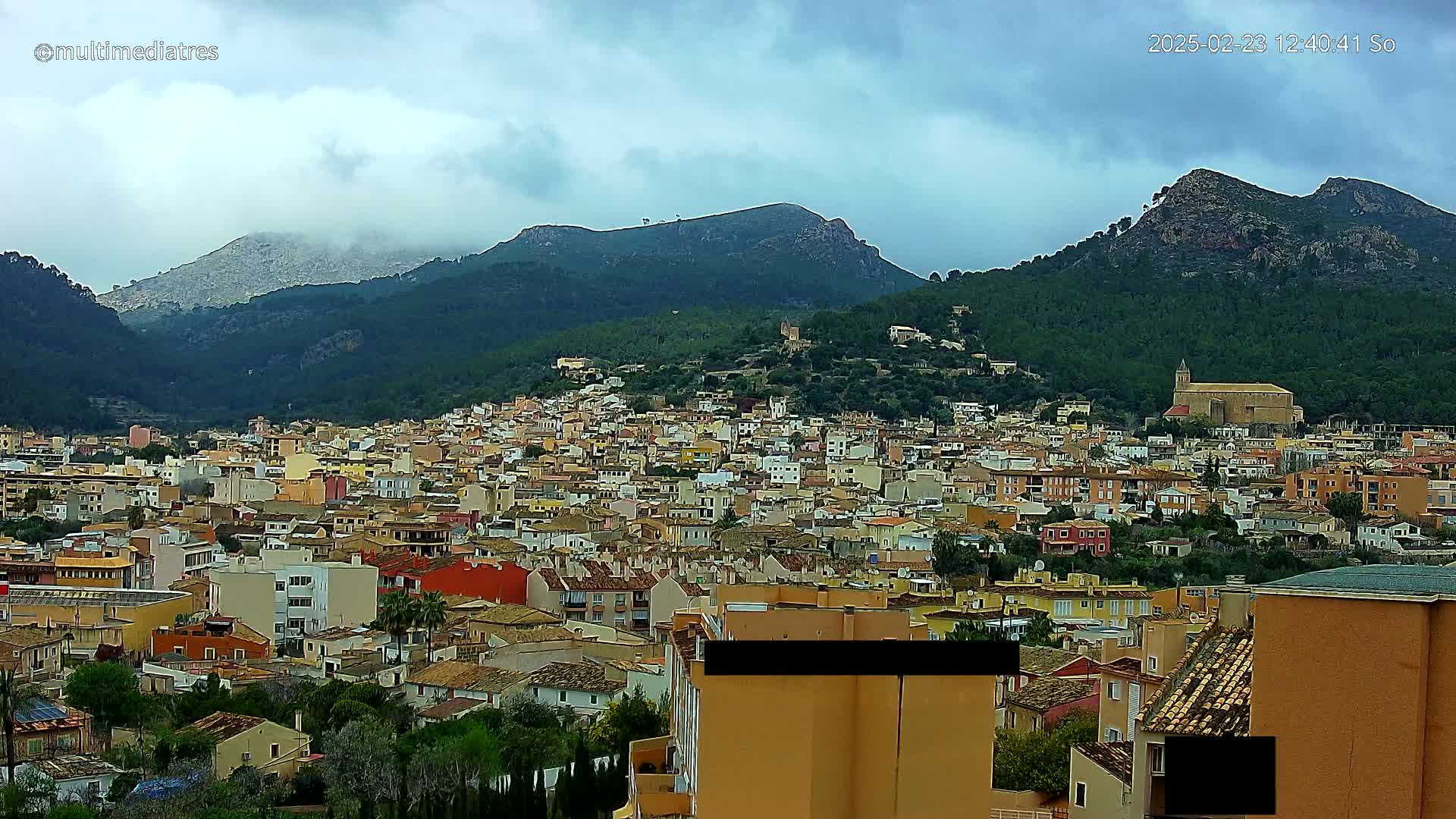 A sprawling town of low-rise buildings nestled in a valley between mountains under a cloudy sky.