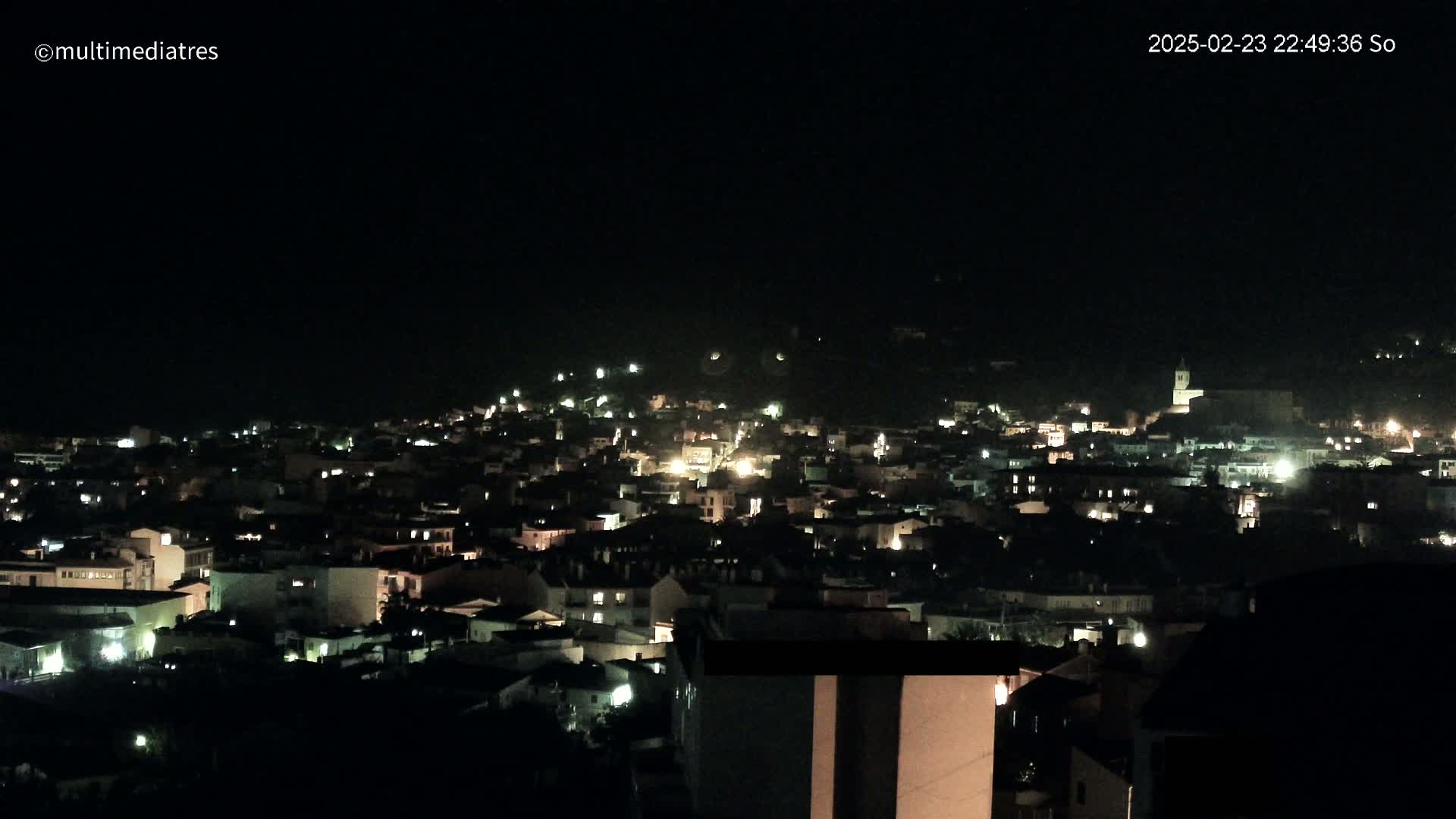 A nighttime, high-angle view shows a town's many illuminated buildings spread across a hillside.