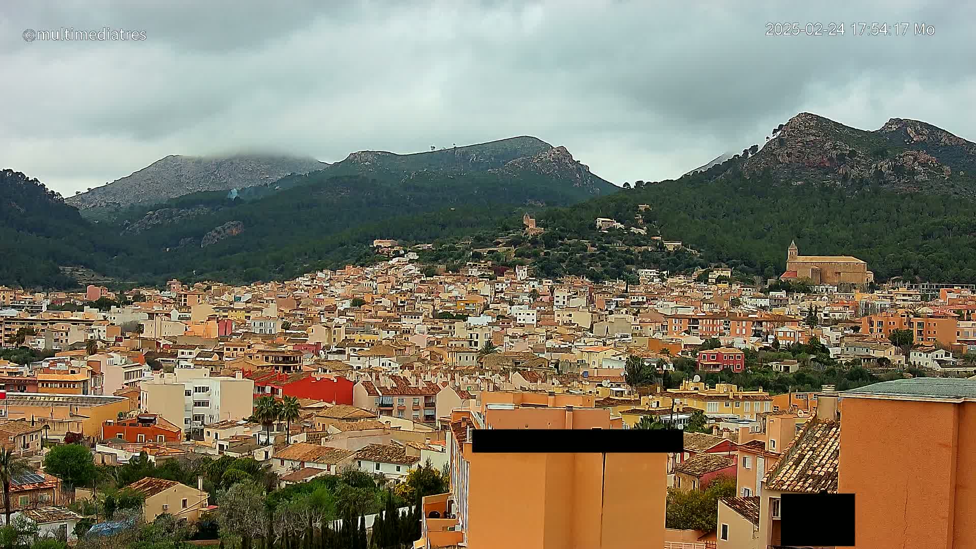 A sprawling town of tan and orange buildings nestled in a valley between green, tree-covered mountains under a cloudy sky.