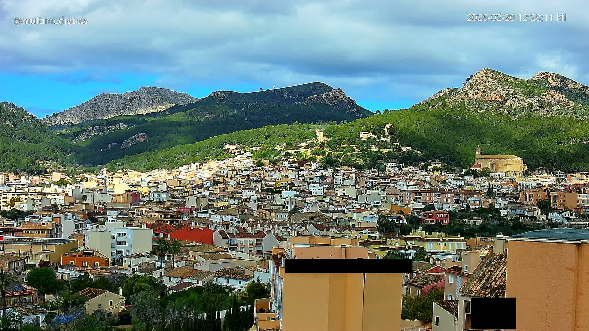 A sprawling town of low-rise buildings nestles at the foot of several verdant, rocky mountains under a partly cloudy sky.