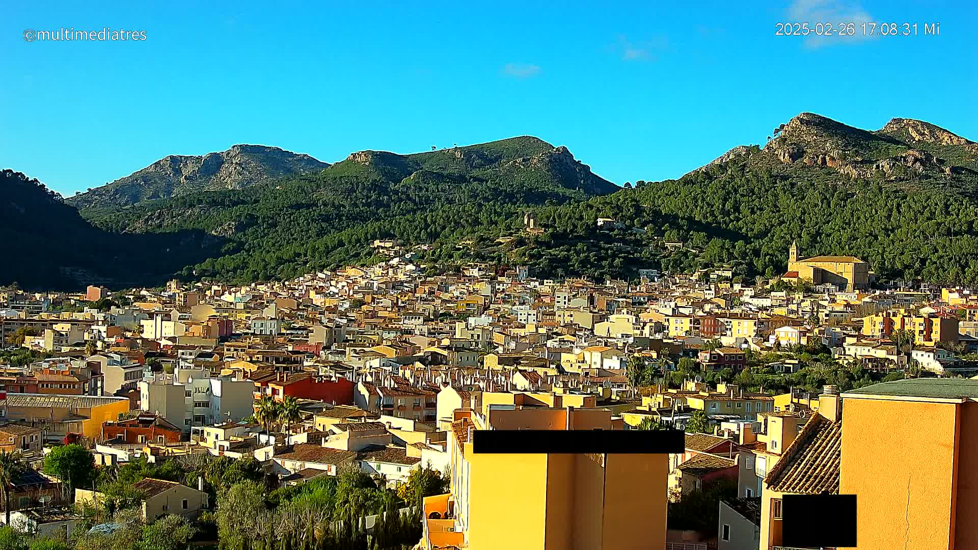 A sprawling town of low-rise buildings nestles at the foot of a verdant mountain range under a clear, blue sky.