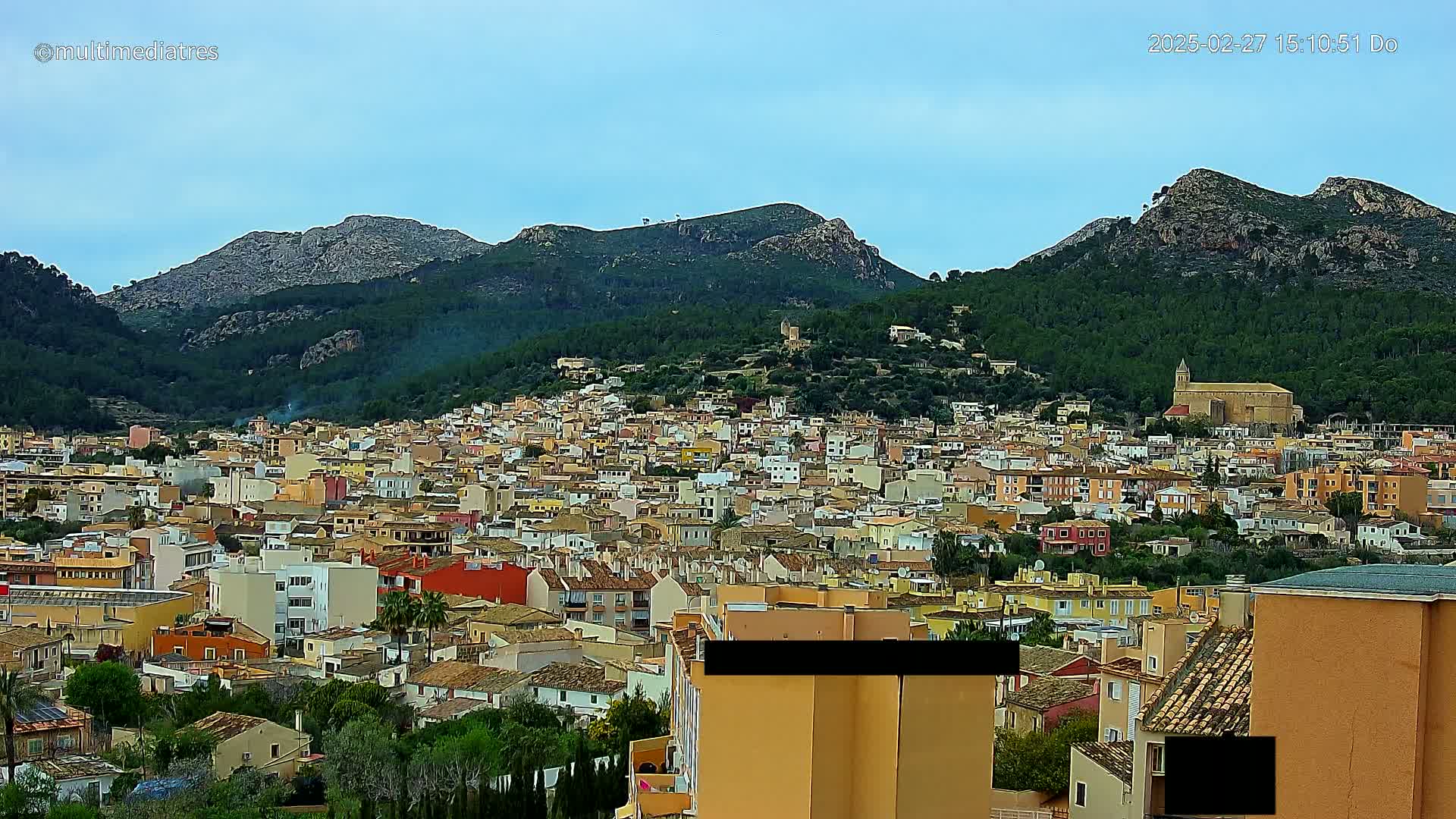 A sprawling town of tan and orange buildings nestled in a valley, surrounded by lush green hills and rocky mountains under a mostly clear sky.