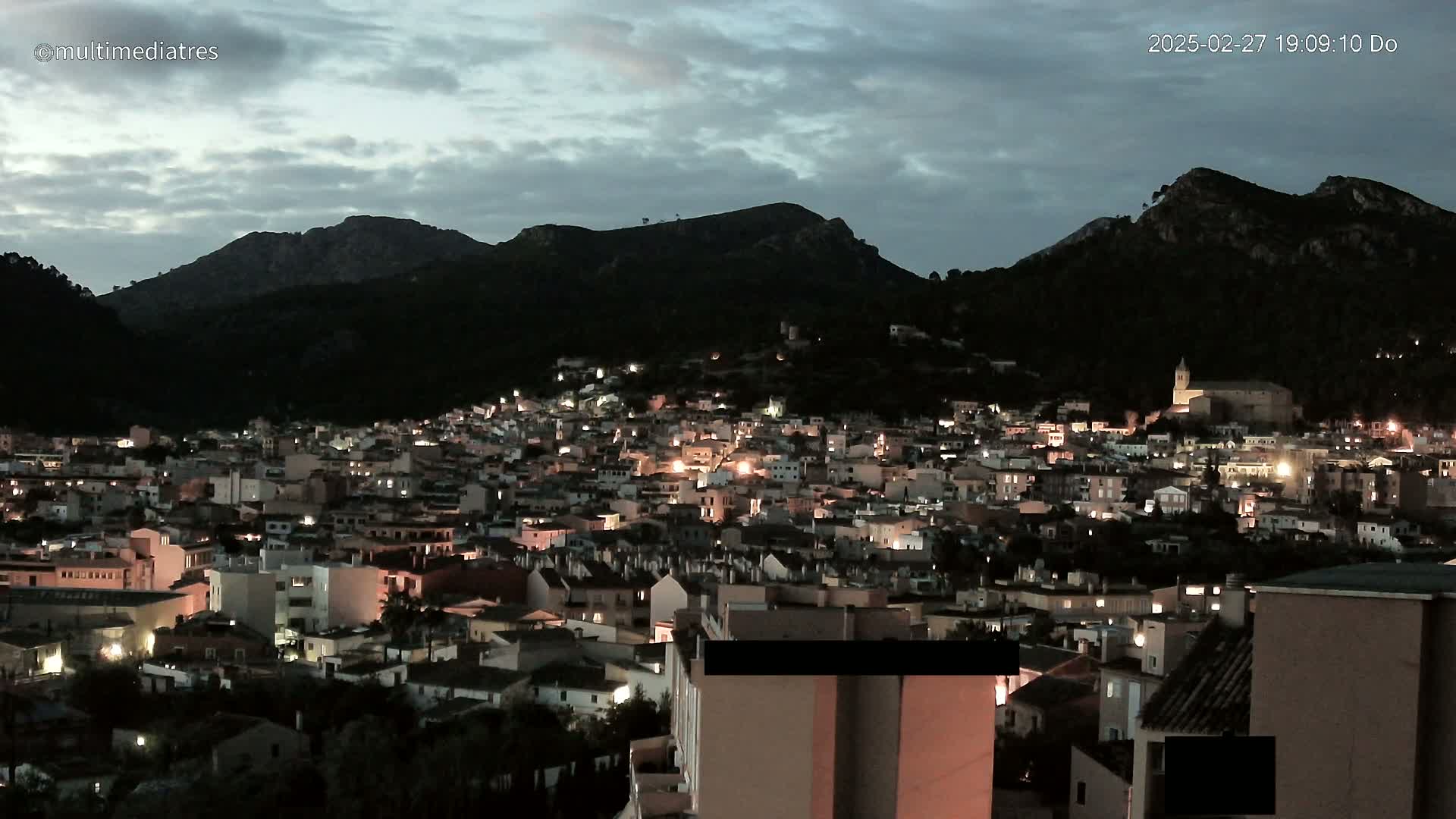 A town nestled in a valley between dark, mountainous hills at twilight under a cloudy sky.