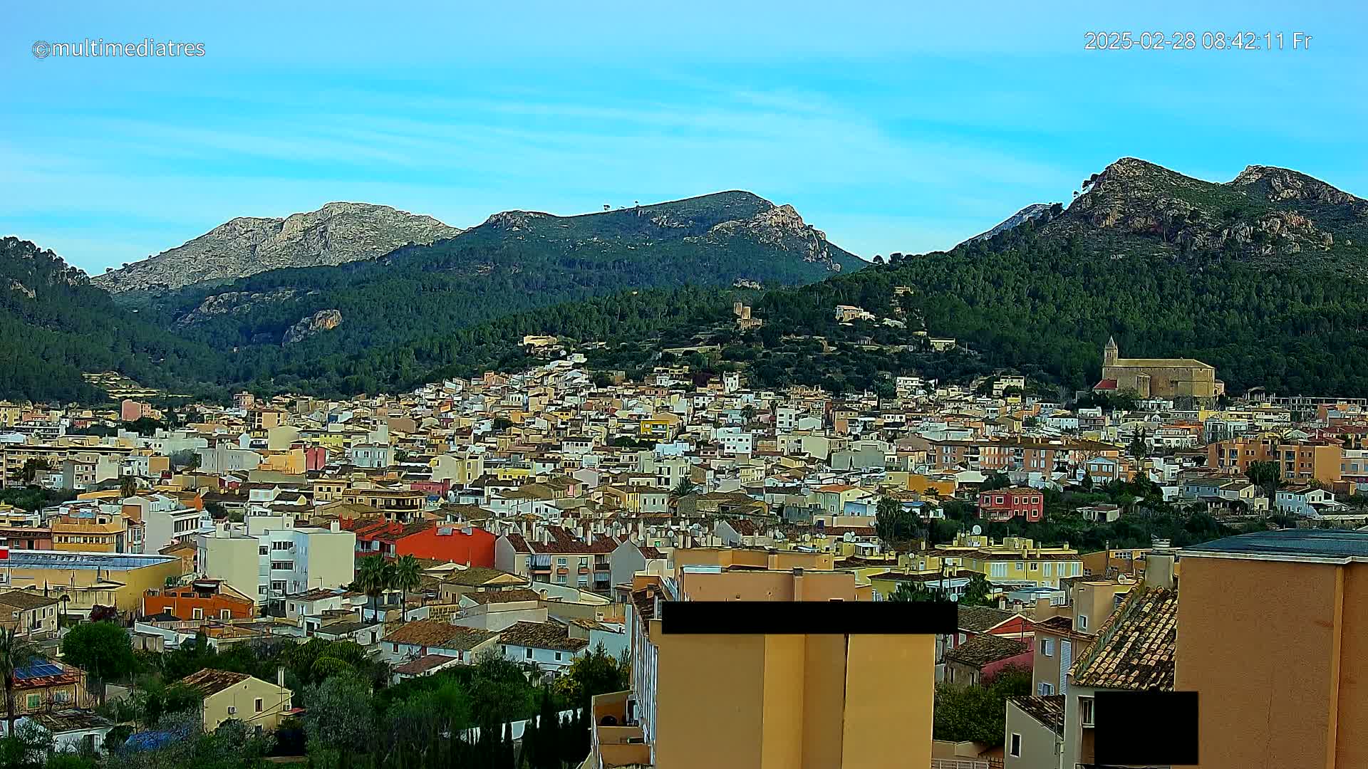 A sprawling town of colorful buildings nestles in a valley surrounded by lush green hills and rocky mountains under a clear blue sky.