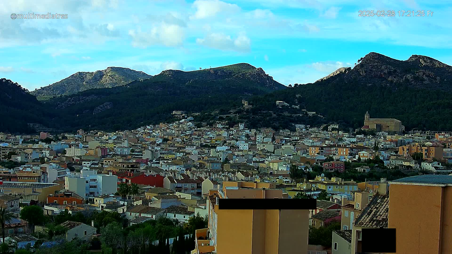 A sprawling town nestled in a valley between verdant mountains under a partly cloudy sky.