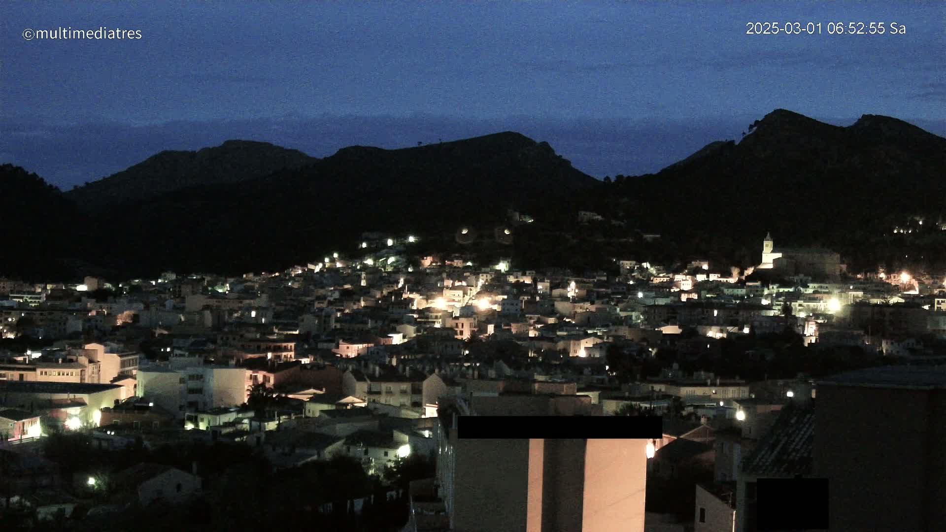 A nighttime view of a town nestled in a valley between dark hills under a twilight sky.